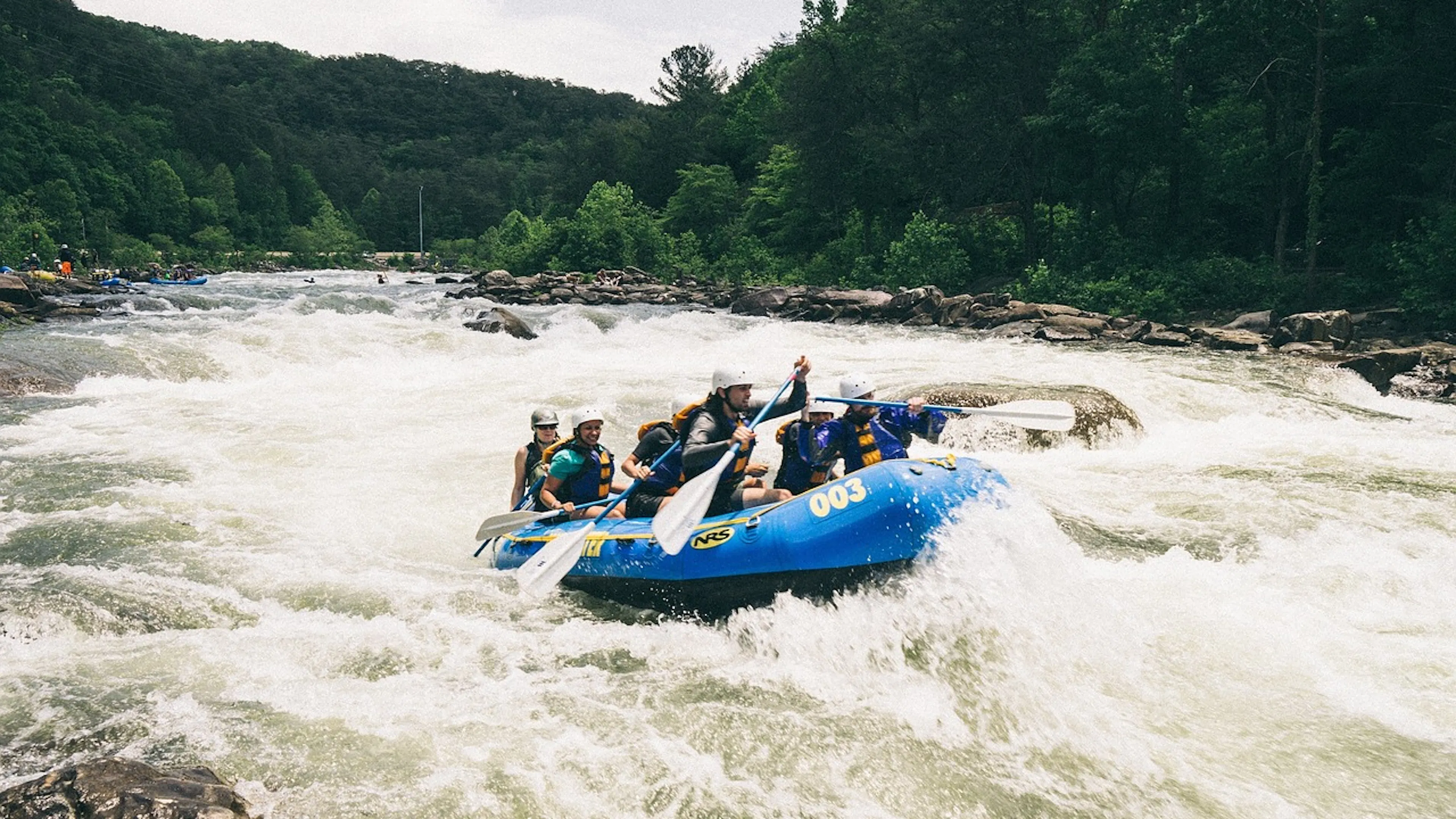 A blue raft with eight people on it paddling through a rapid on a river