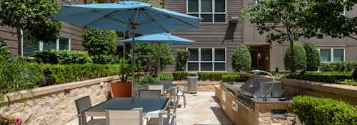 AMLI River Oaks outdoor grilling and dining area with a gas grill surrounded by lush greenery and shaded by blue umbrellas