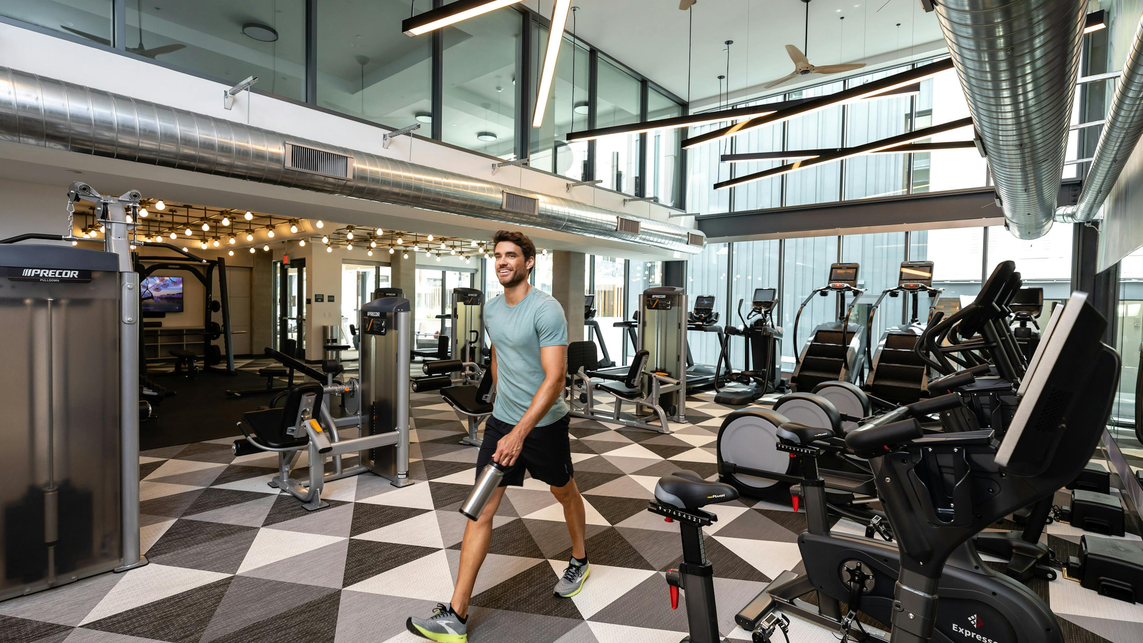 Smiling young man carrying a water bottle walking through the fitness center by the cardio machines at AMLI Old Pasadena