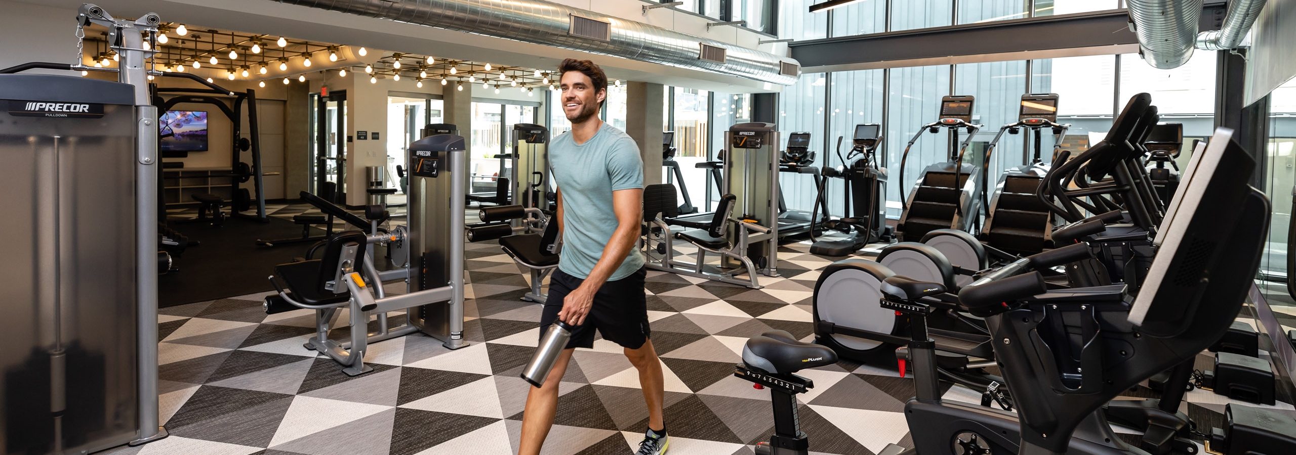 Smiling young man carrying a water bottle walking through the fitness center by the cardio machines at AMLI Old Pasadena