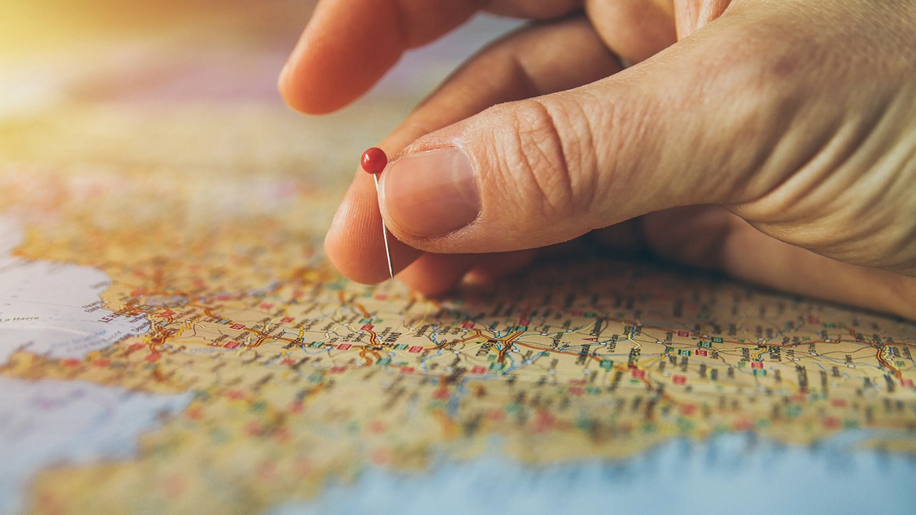 Extreme close up of a hand with the thumb and middle finger about to poke a red pin into an old road map