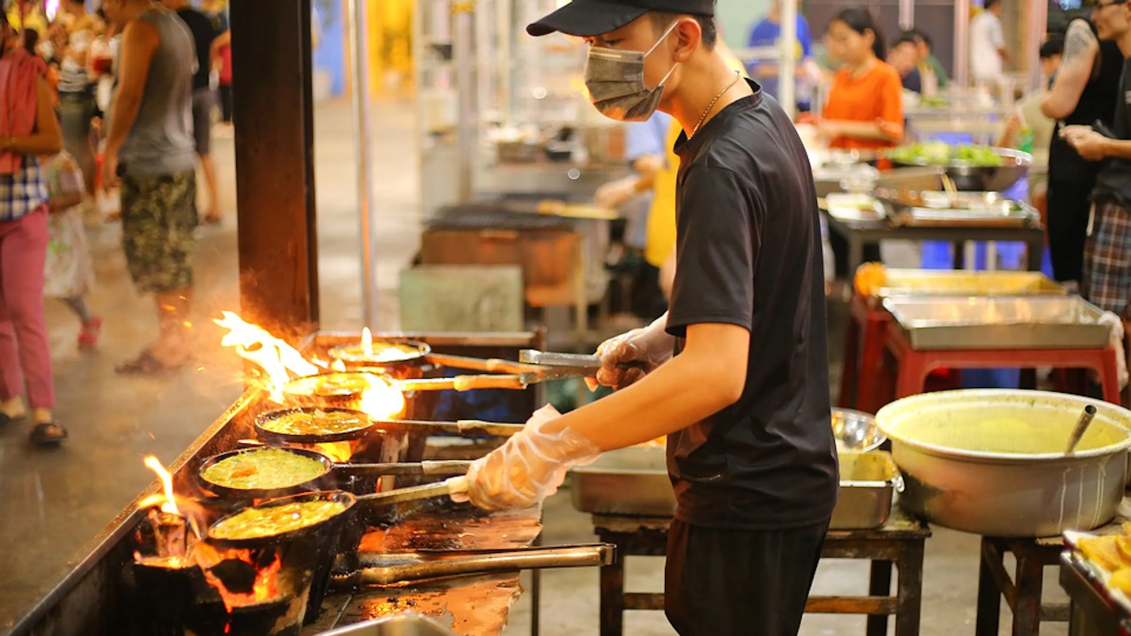 A person cooking street food with six different pots over a gas grill 