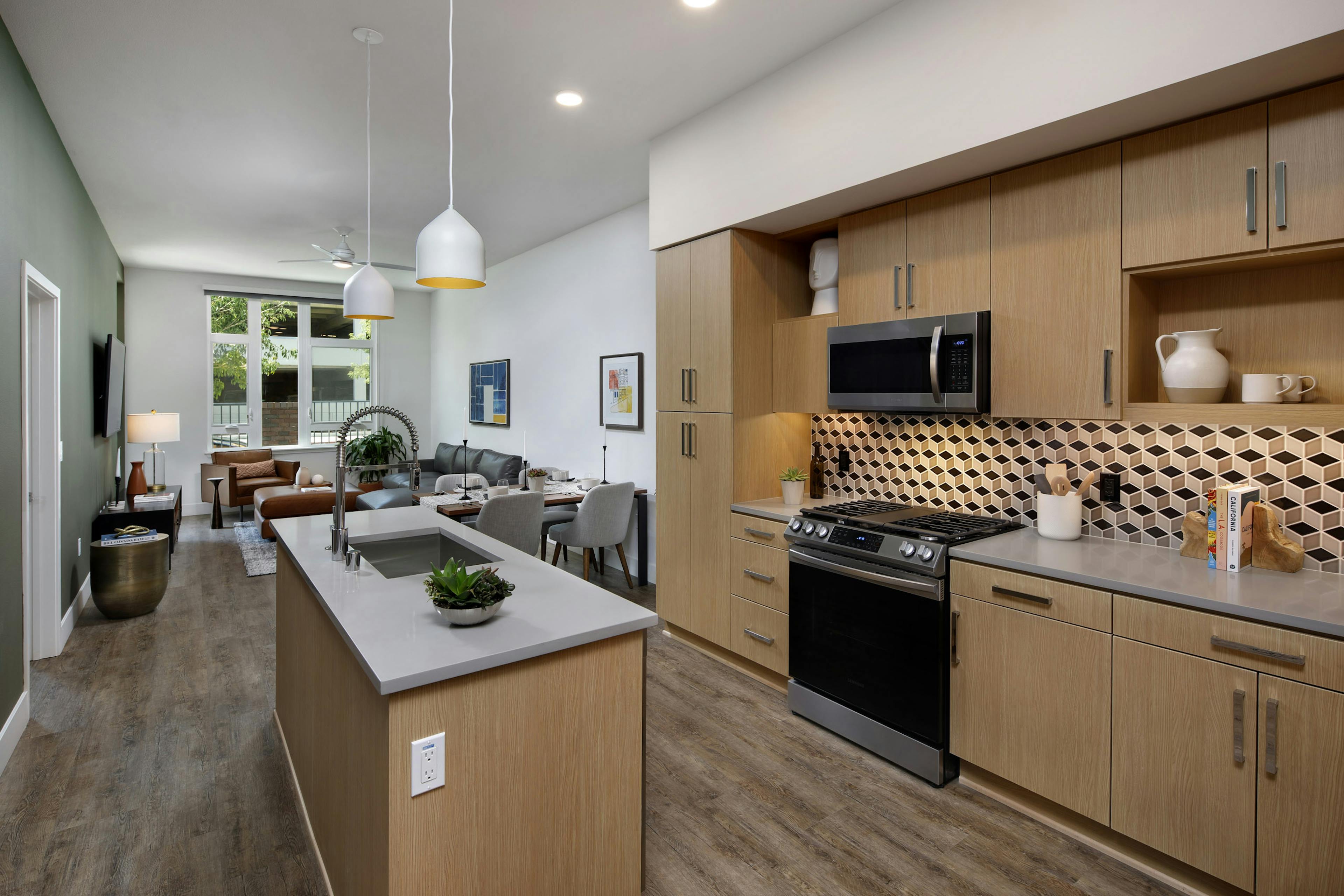 Interior of AMLI Old Pasadena apartment kitchen with light cabinets, and checkered backsplash facing spacious living room