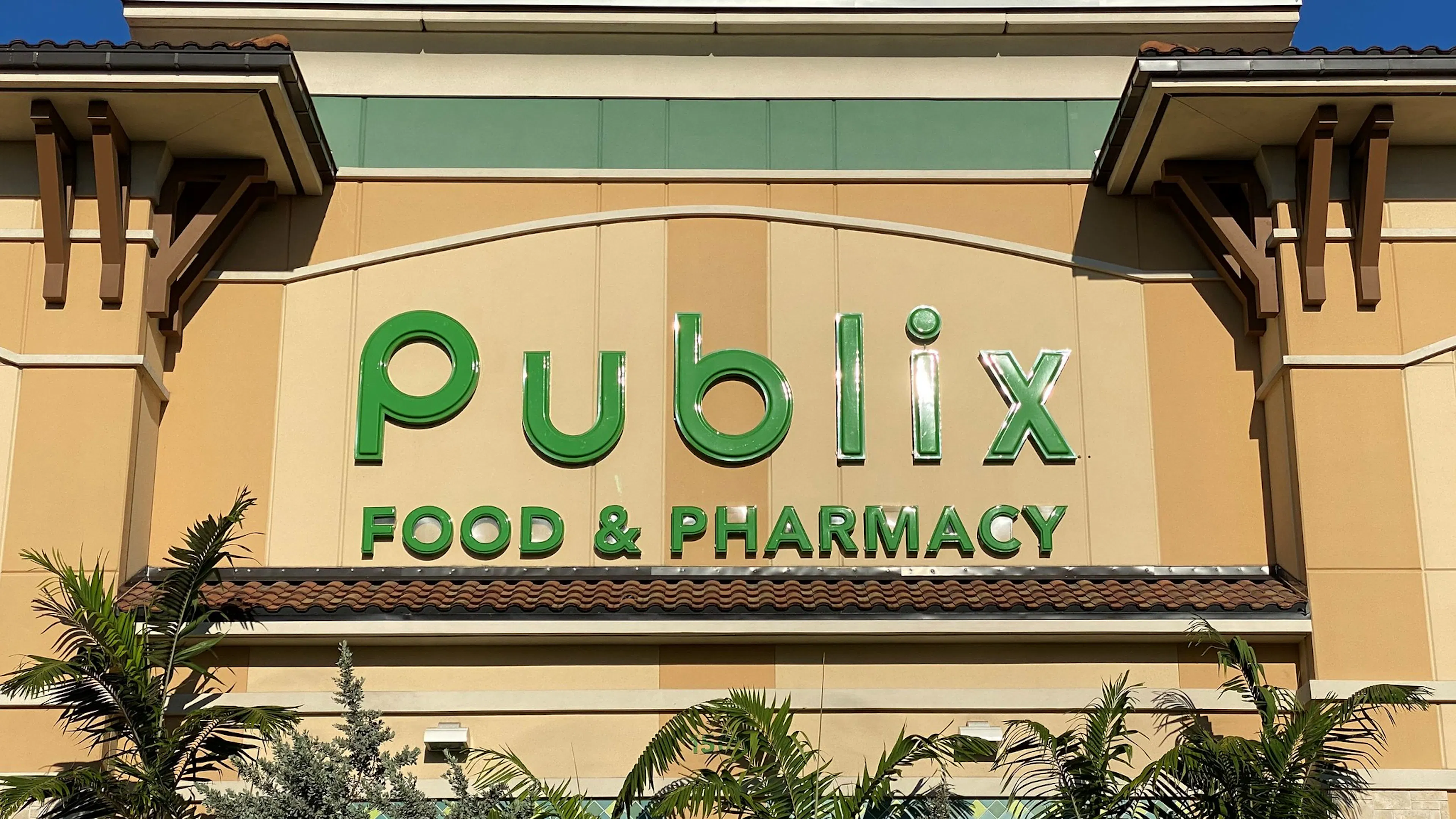 The Publix logo against a stucco exterior and a few palm trees beneath