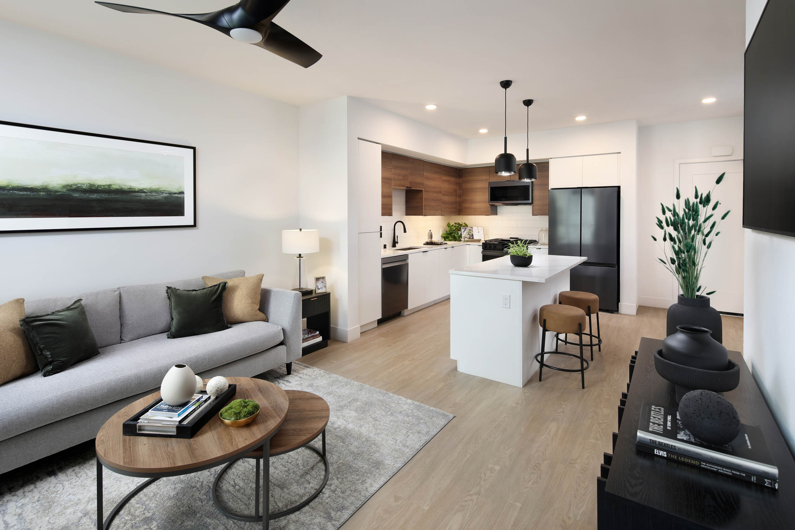 Open-concept living room and kitchen with gray sofa, round coffee table, white island, bar stools, wood cabinets, pendant lights, and TV.