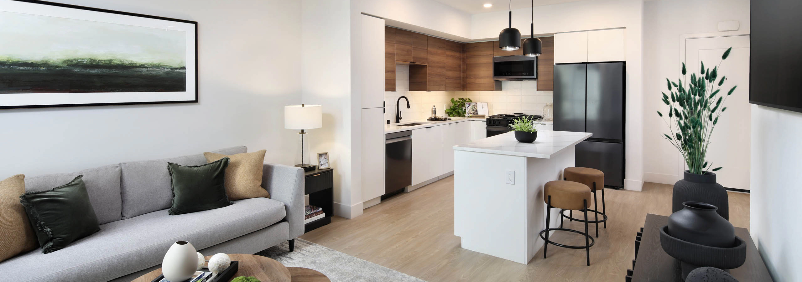 Open-concept living room and kitchen with gray sofa, round coffee table, white island, bar stools, wood cabinets, pendant lights, and TV.