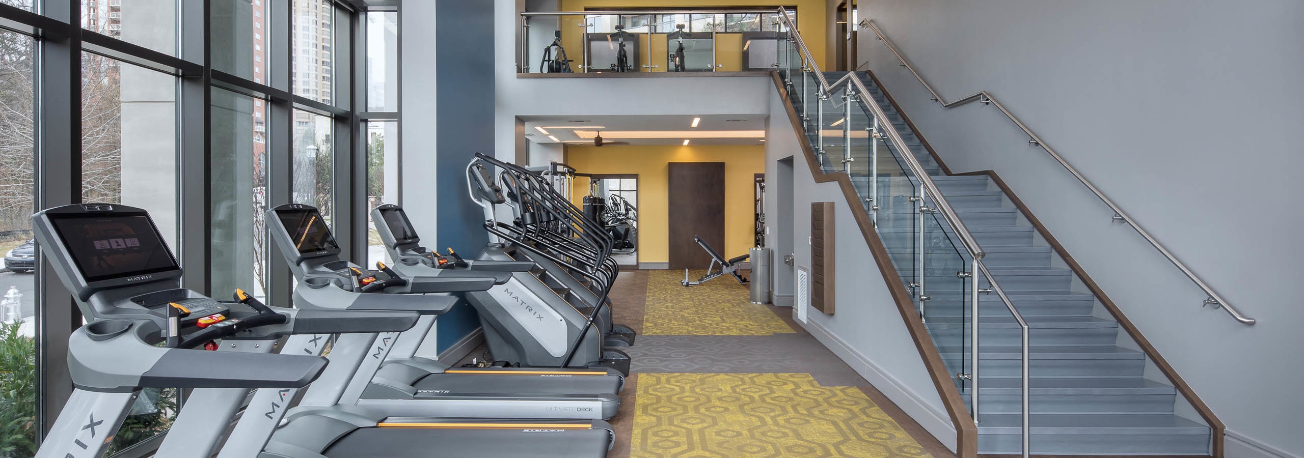 Fitness center at AMLI 3464 with yellow carpet and treadmills near tall windows with a modern staircase in the background