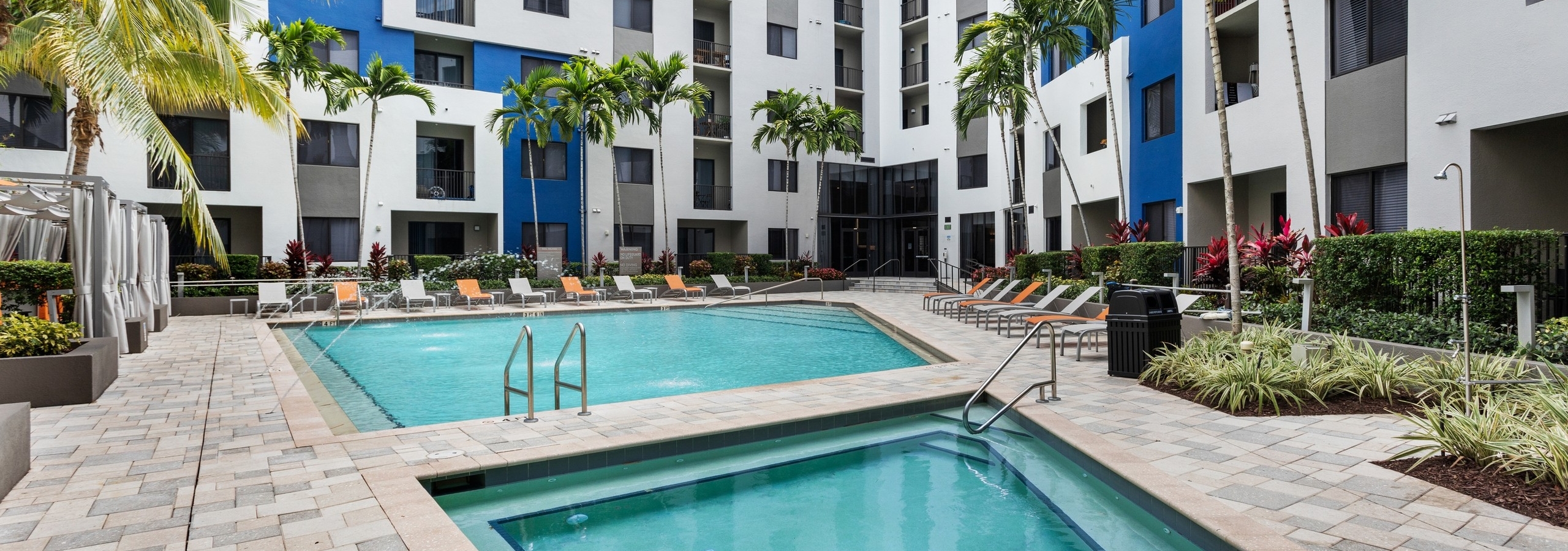 The pool at AMLI 8800 with water feature over pool and adjacent spa with gray and orange lounge chairs and cabanas