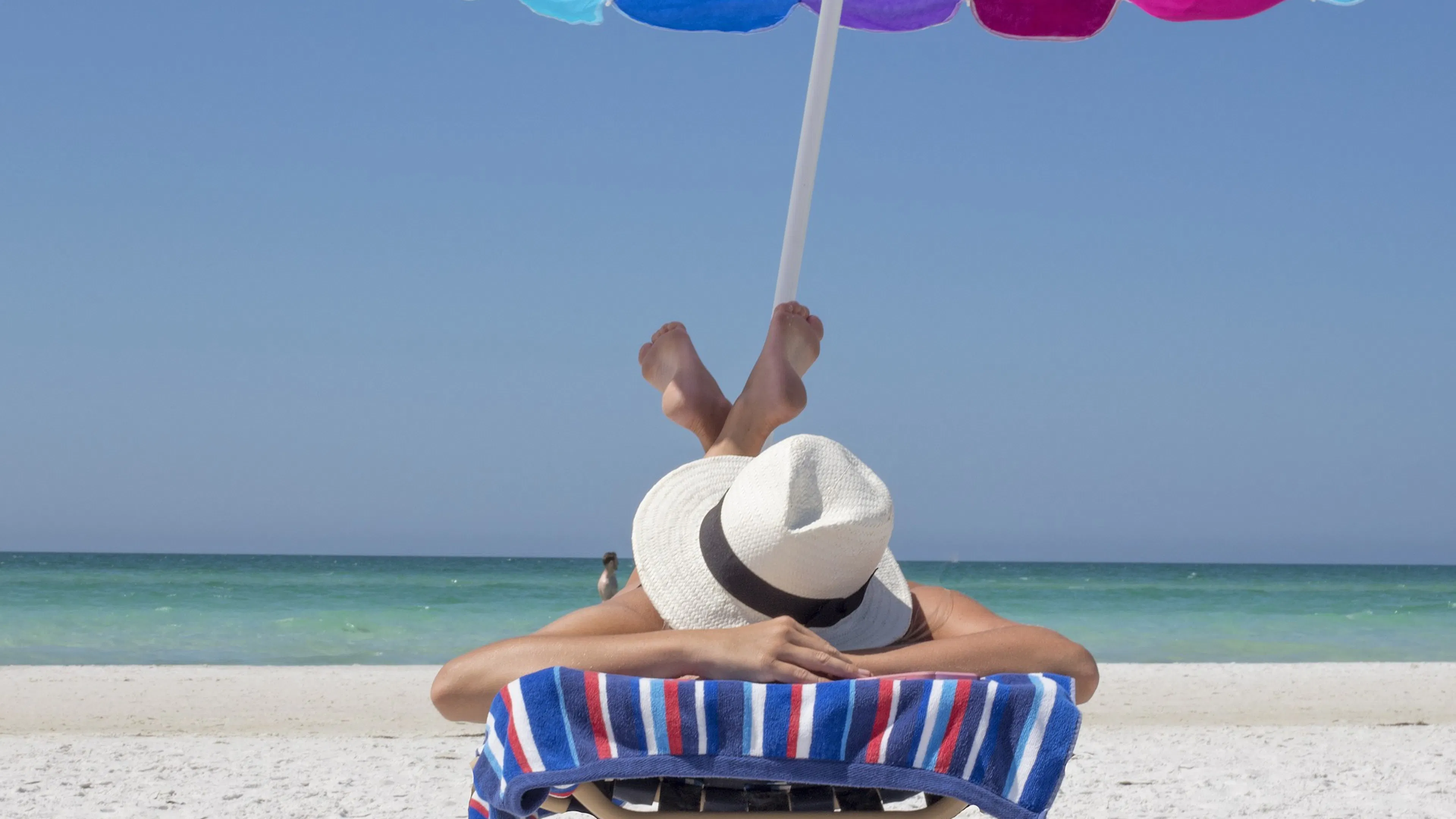 A woman in a white hat lying face down on a beach chair under an umbrella with the ocean in the background