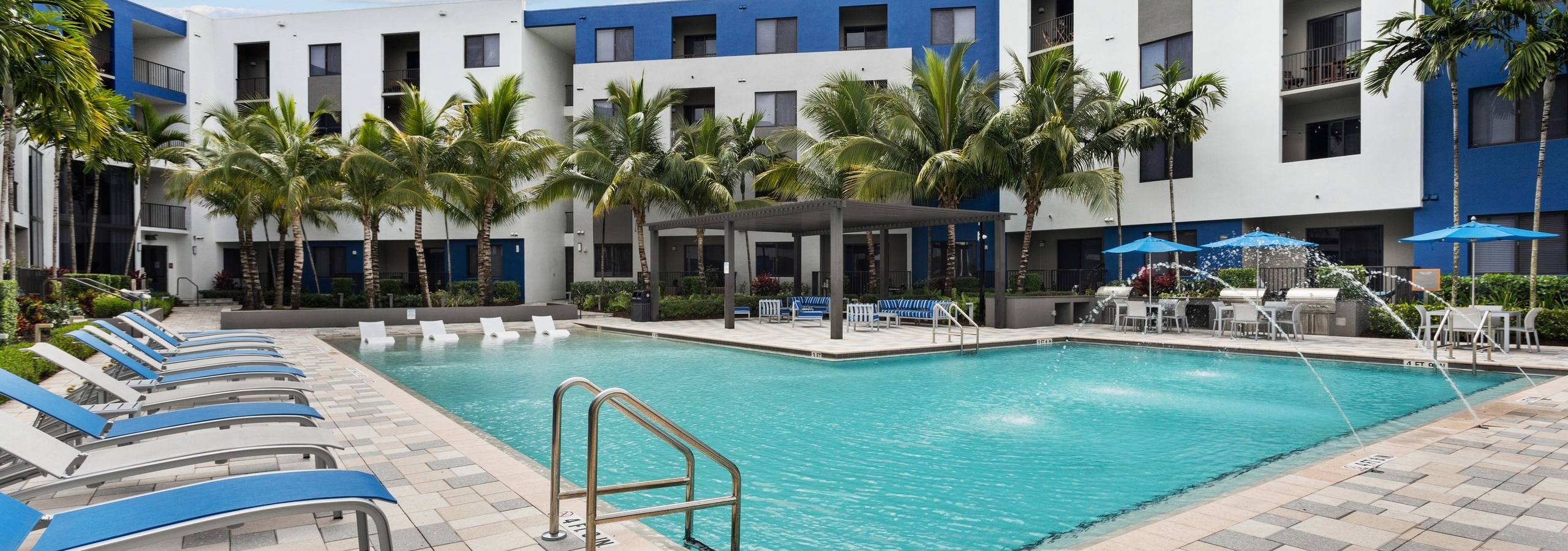 AMLI 8800 pool with water feature and blue and gray lounge chairs a pergola with seating and table and chairs with umbrellas