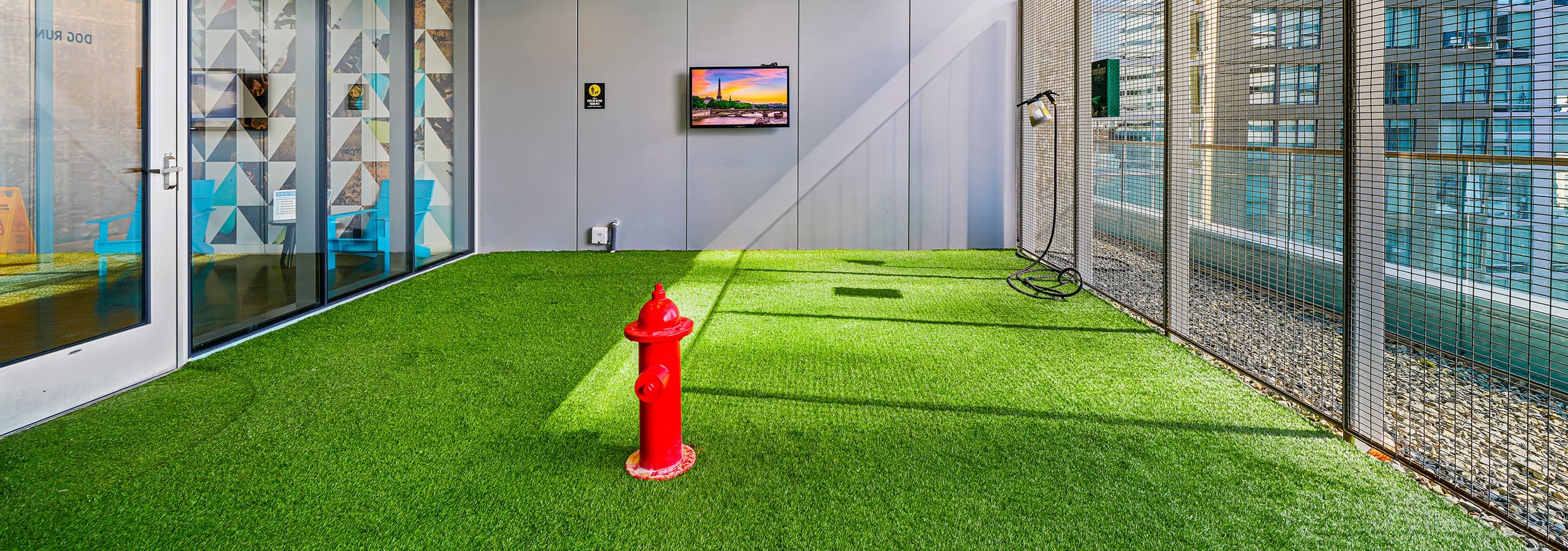 Pet park at AMLI Arc apartment building with fire hydrant in center of green turf and fenced wall and TV on gray wall