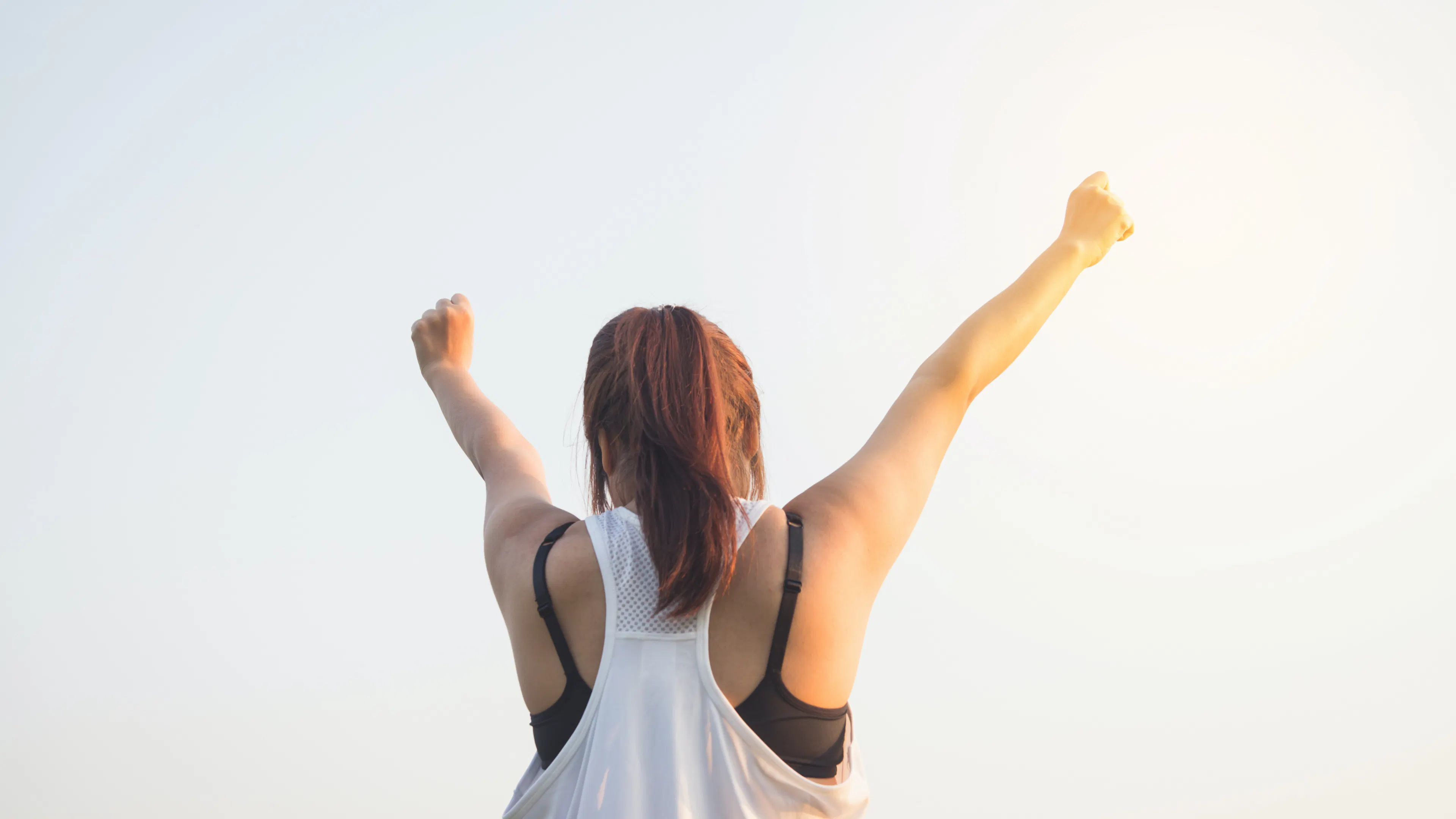 View of the back of a woman in a white jogging tank top and pony tail top raising her arms over her head 