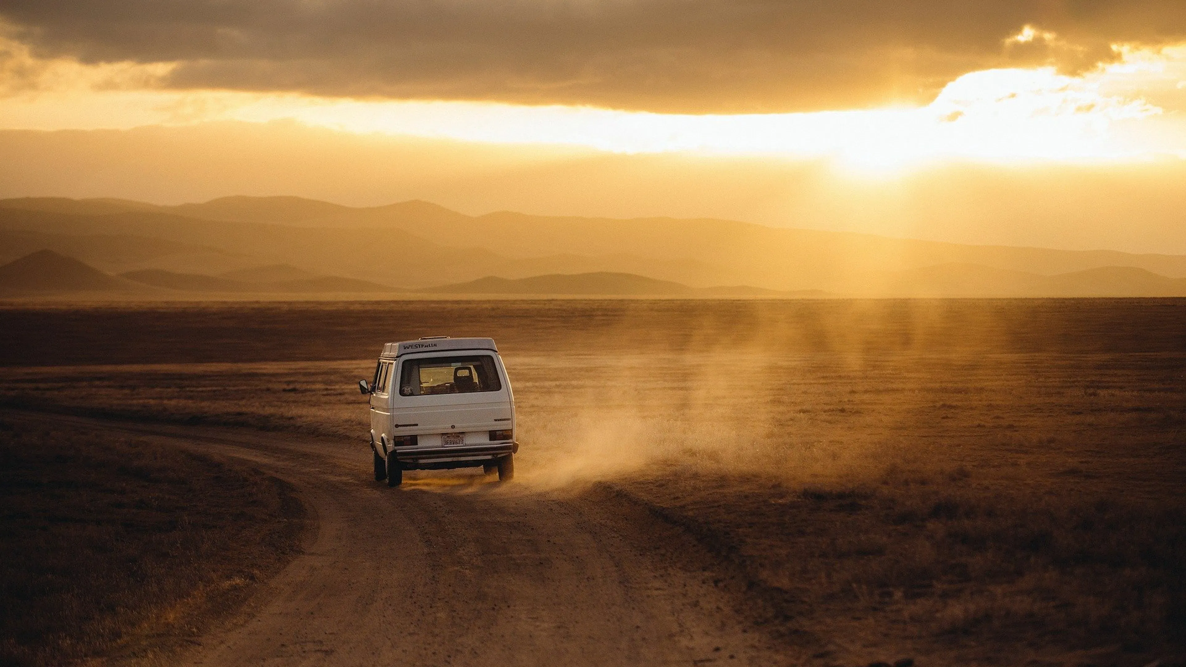 An old Volkswagen van kicking up dust on a rural back road in the sunset