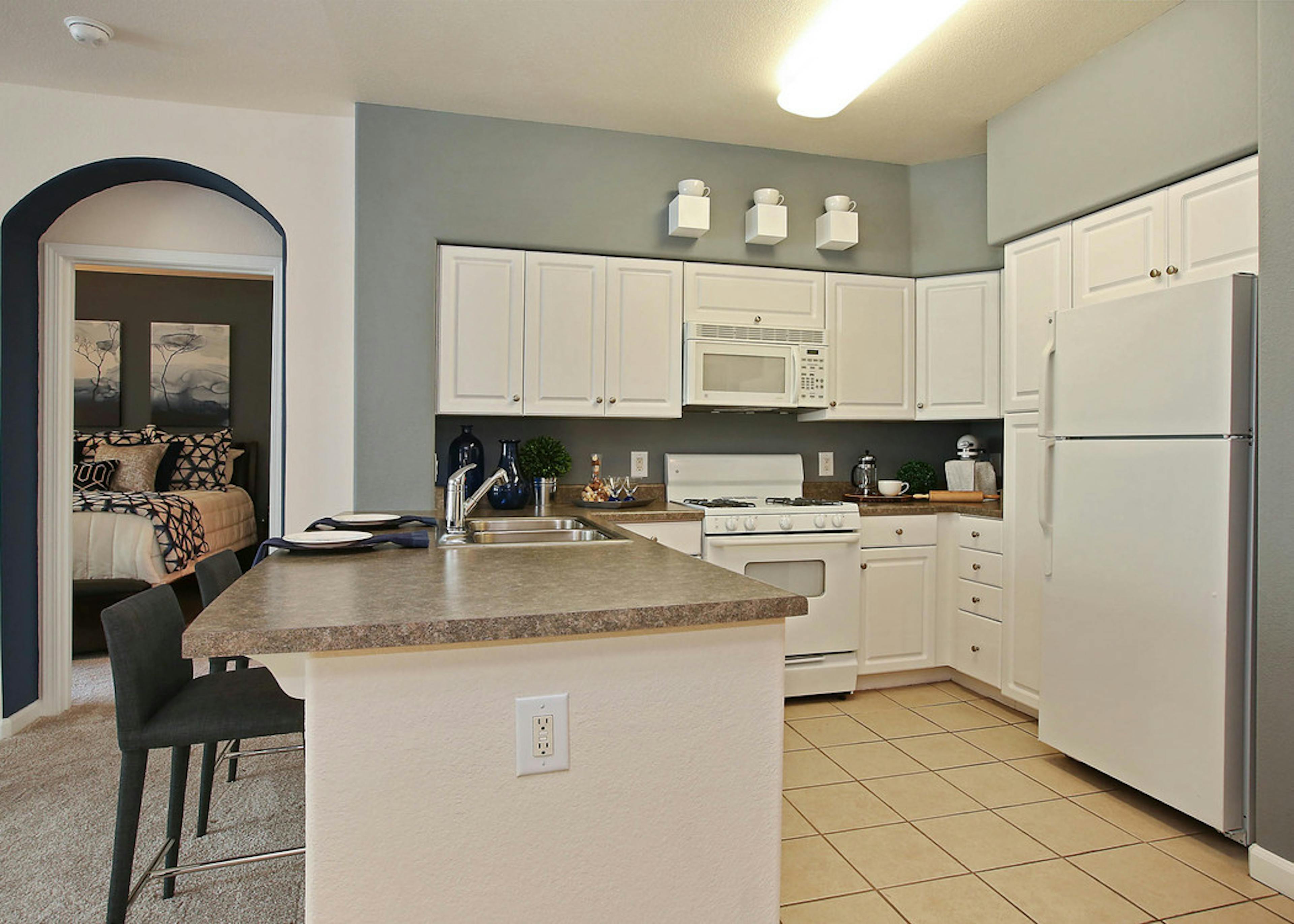 Interior of AMLI Warner Center apartment kitchen with white cabinetry, double sink and white appliances and peek into bedroom