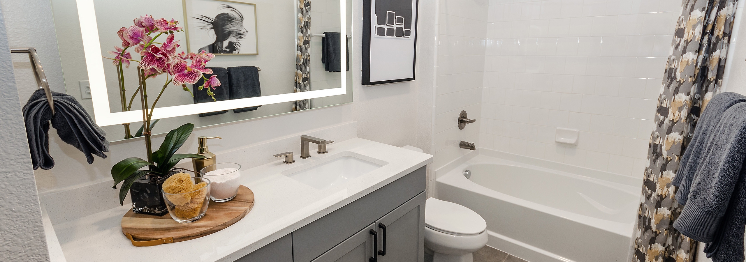 AMLI Quadrangle bathroom with a grey wood vanity sink with a white countertop next to a toilet and a white tiled shower tub
