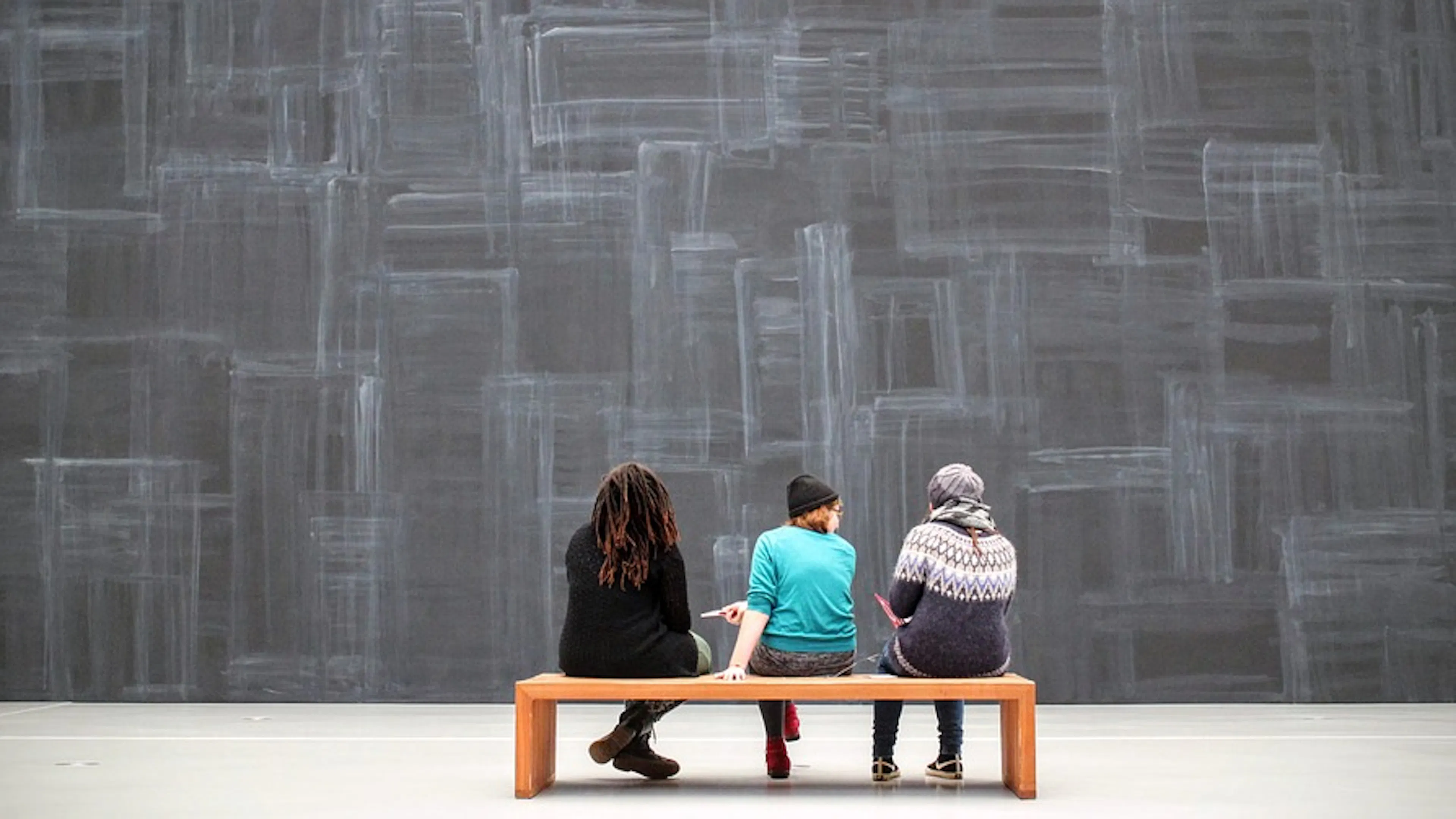 Three people sitting on a low bench facing a large grey concrete wall