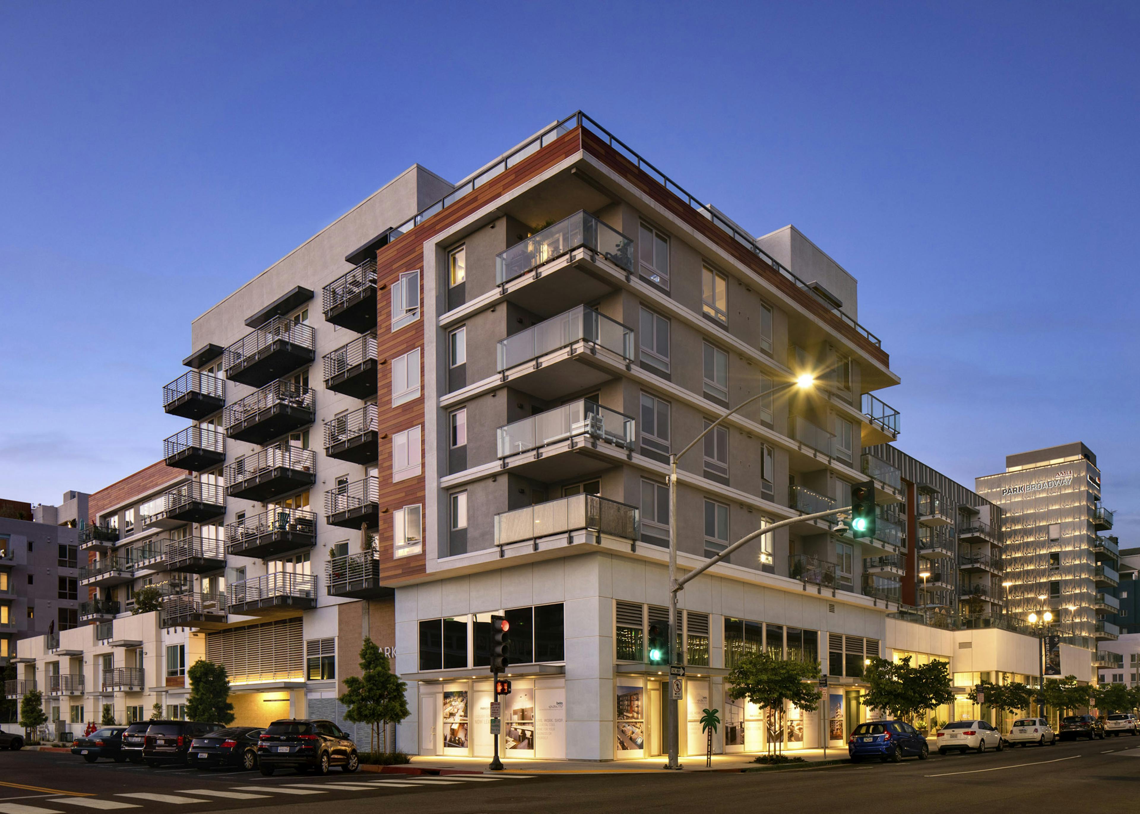 Corner view of AMLI Park Broadway apartments with lit up first floor retail spaces with cars and trees lining the streets