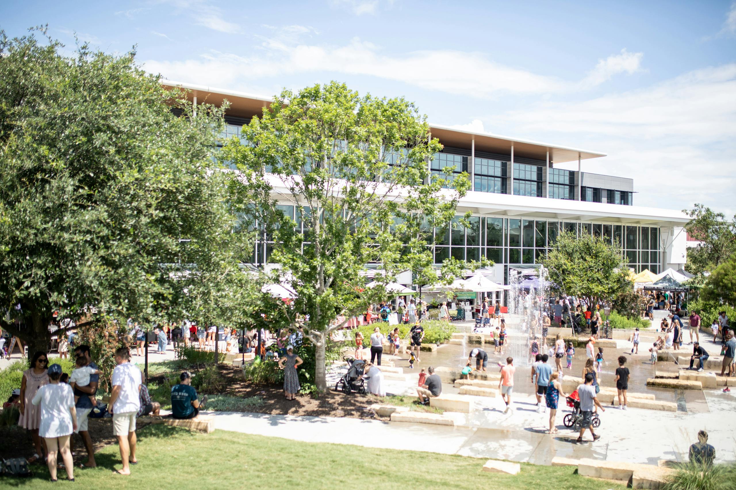 People playing in big fountain in front of Mueller farmers market alongside glass building with large trees on a sunny day