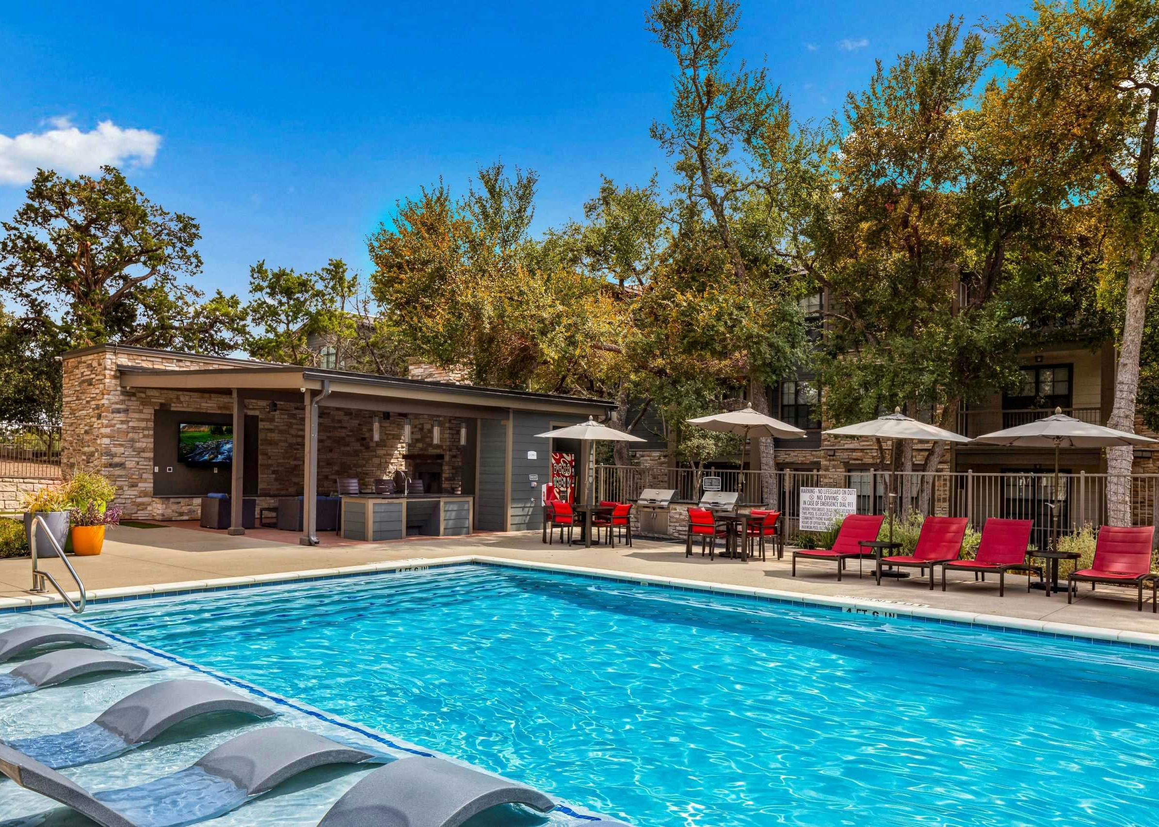 View of pool at AMLI Covered Bridge with in pool lounge chairs and red chairs with taupe umbrellas with stacked stone cabana
