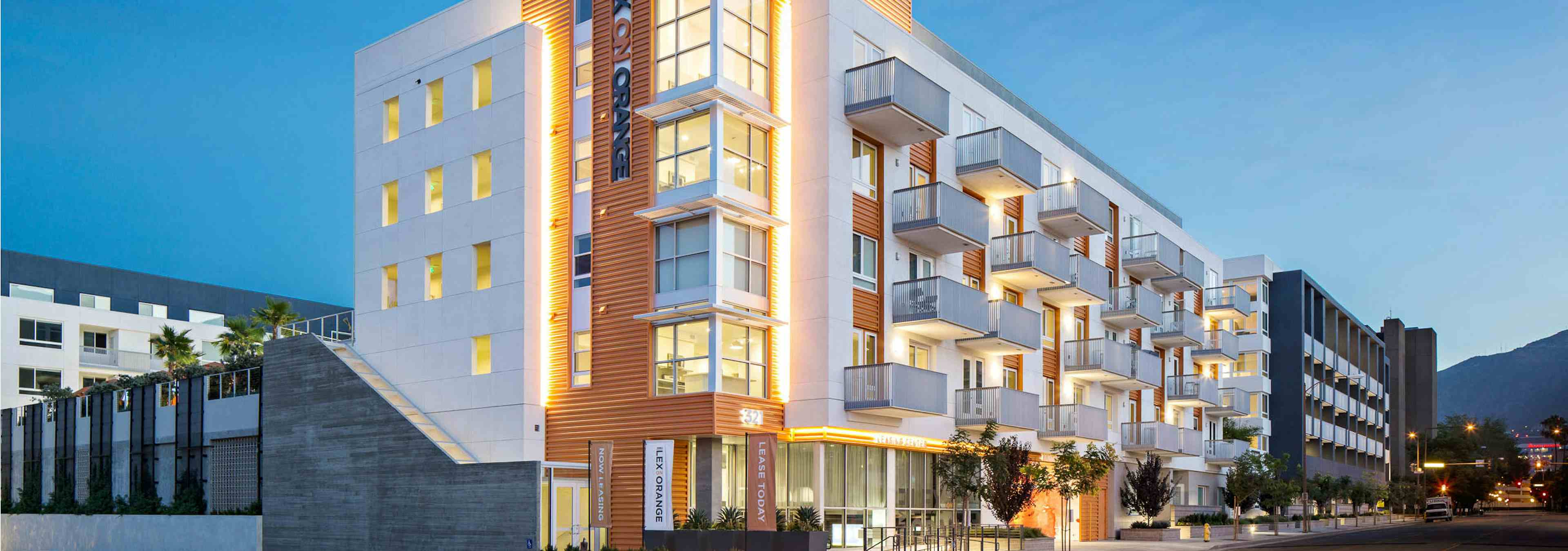 Exterior orange, white and blue rendering of AMLI Lex on Orange apartment building lit up at dusk and trees on empty sidewalk