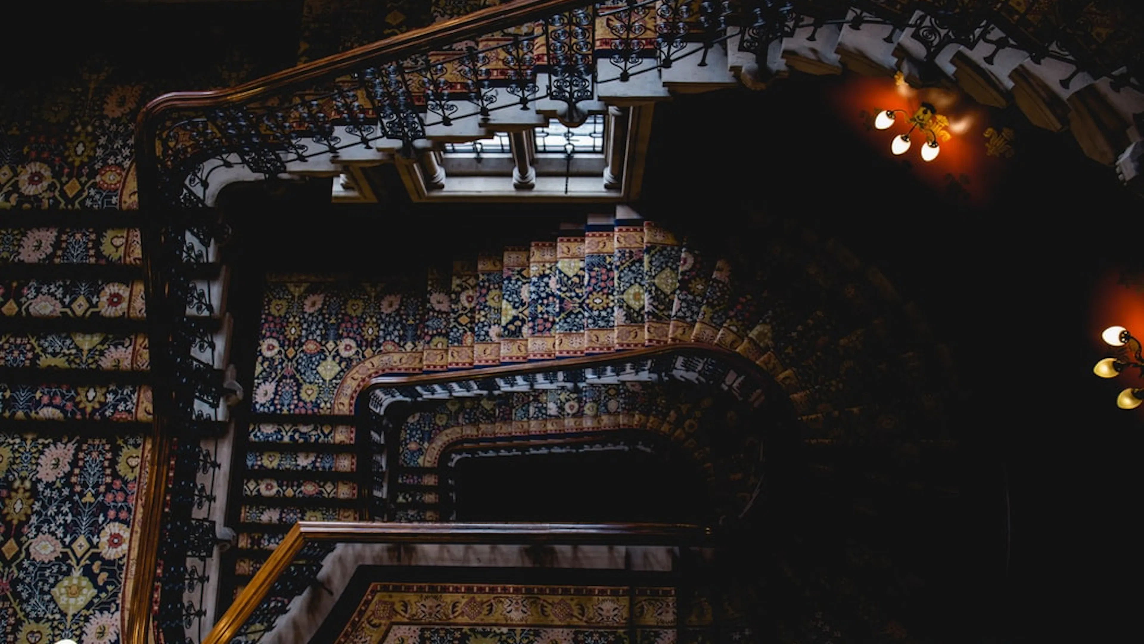 An overhead view of a dimly lit wooden staircase with elaborate carpet and dim lamps on the walls spiraling down 