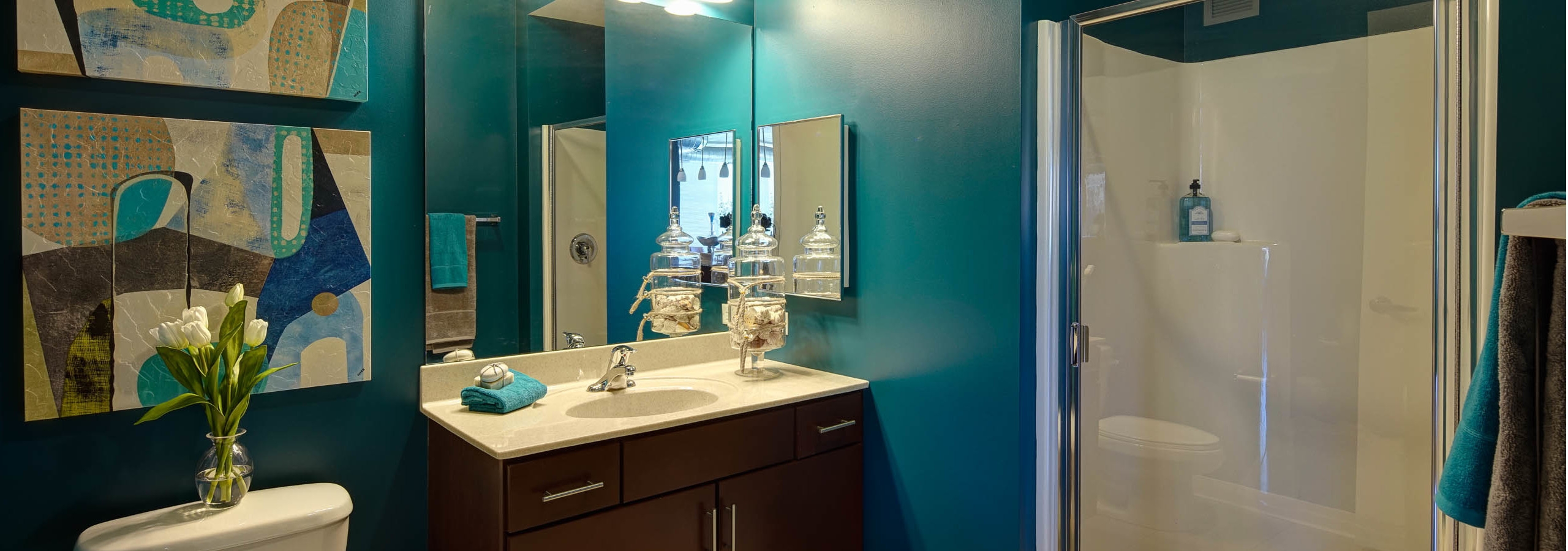 An AMLI 900 bathroom with geometric canvases hanging on teal walls featuring a white toilet, bathtub and dark wood vanity