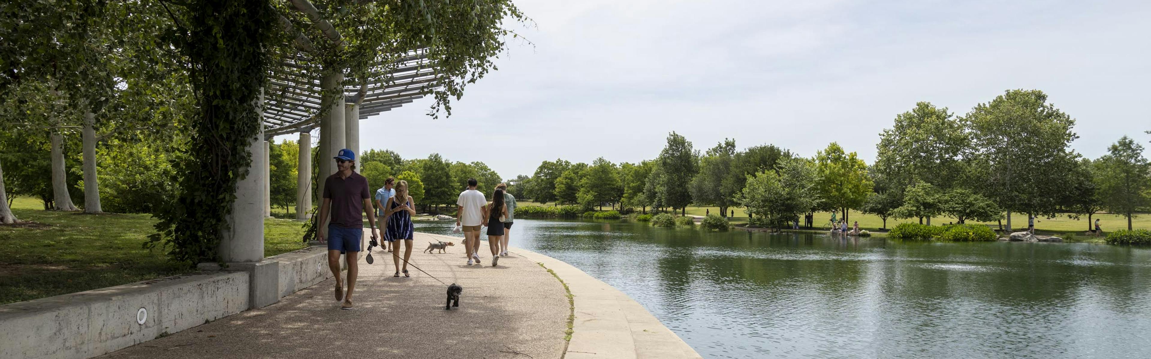 Daytime view of Mueller Lake featuring Mueller Lake Park visitors walking their dogs under a white shaded pavilion