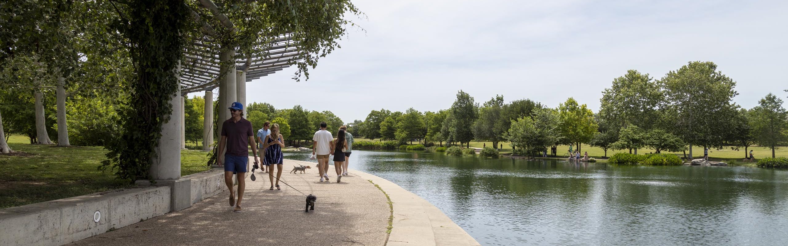 Daytime view of Mueller Lake featuring Mueller Lake Park visitors walking their dogs under a white shaded pavilion