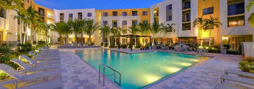One of two swimming pools at AMLI 8800 at dusk surrounded by lush palm trees lit up, lounge chairs and building exterior