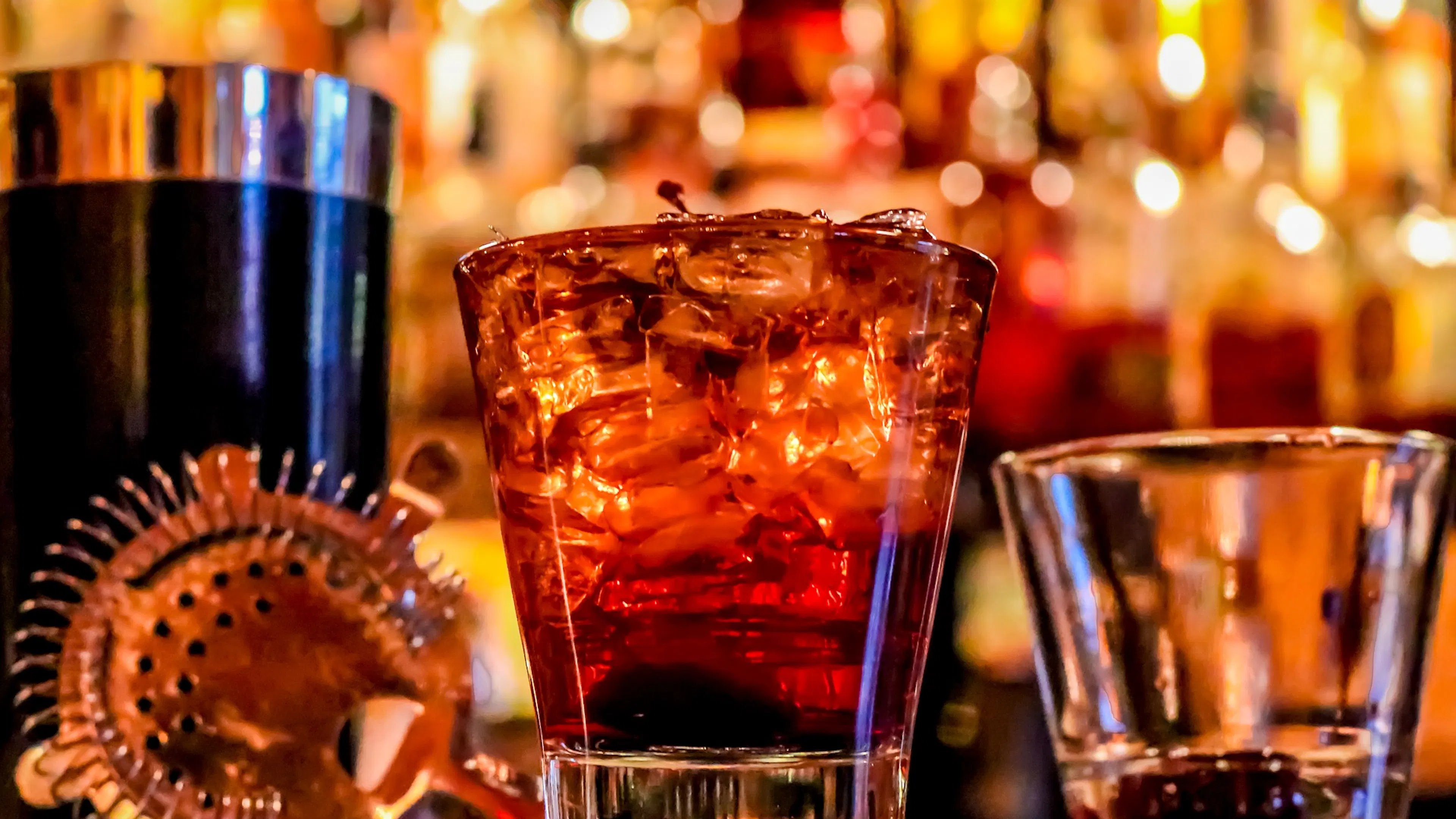 Two rocks glasses next to a cocktail shaker on a bar with brightly lit bottles behind them