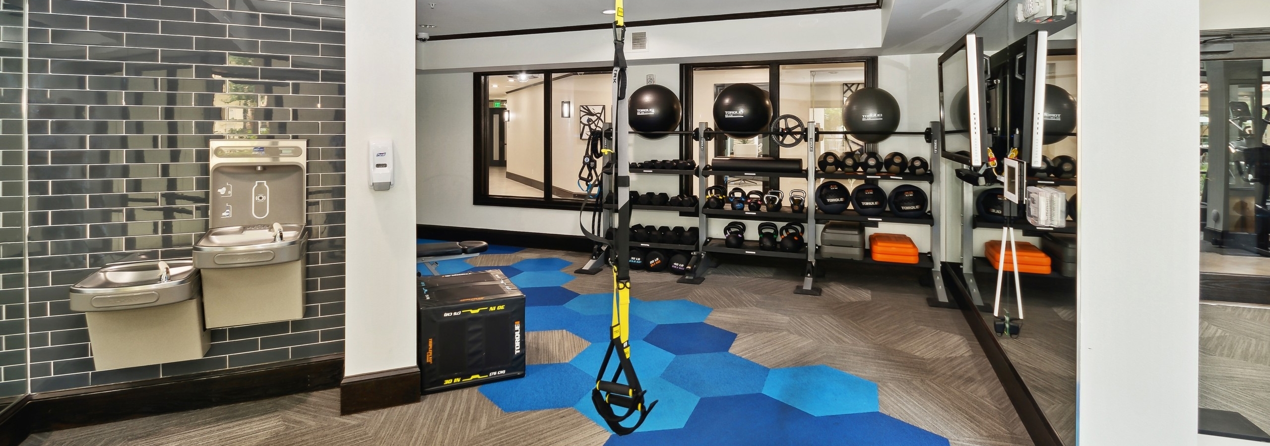 Fitness center at AMLI Campion Trail with weights on shelves and drinking fountains and and blue geometric pattern on floor
