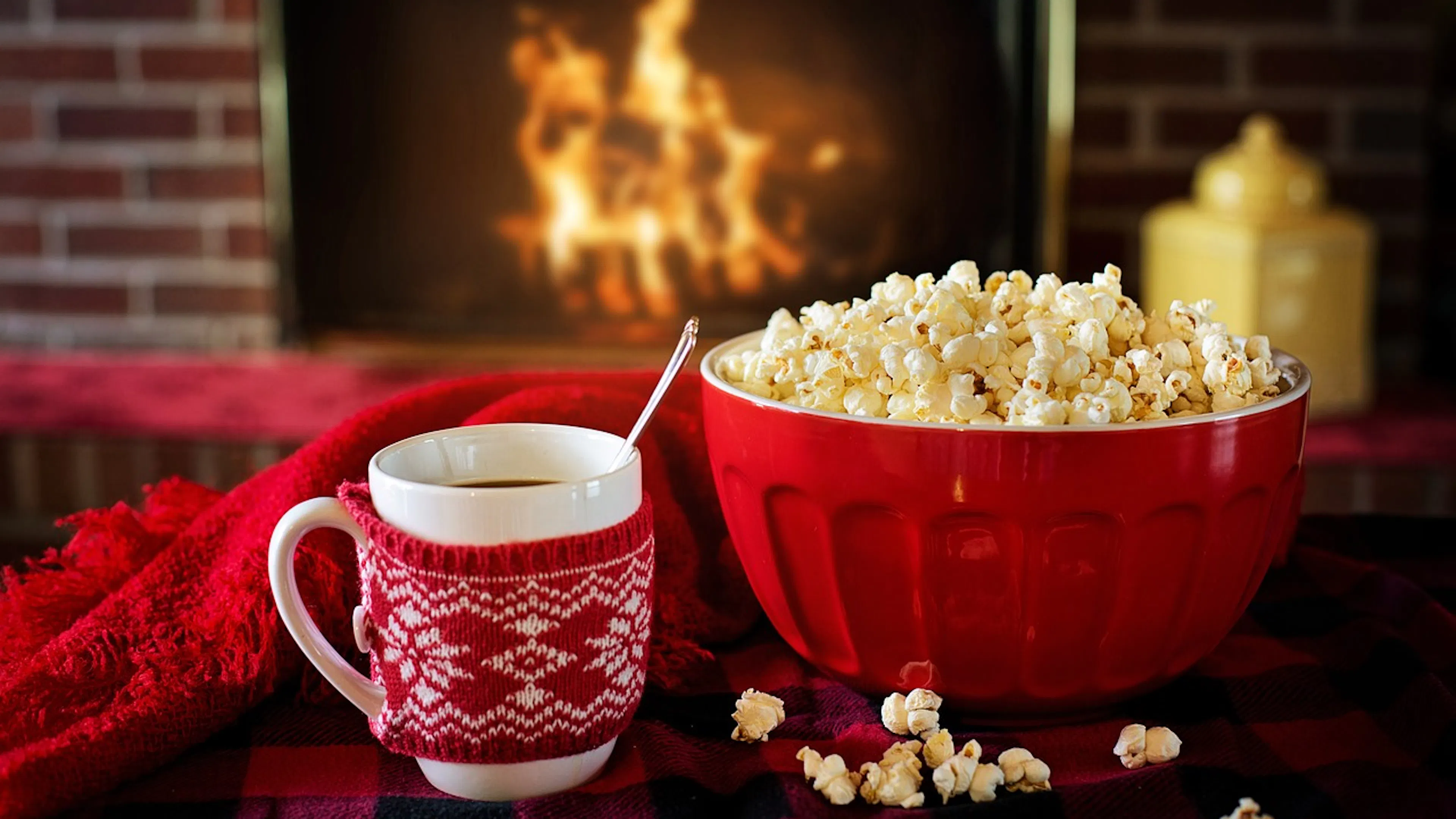 mug with red sweater cozy and red bowl with popcorn in front of fire with red scarf