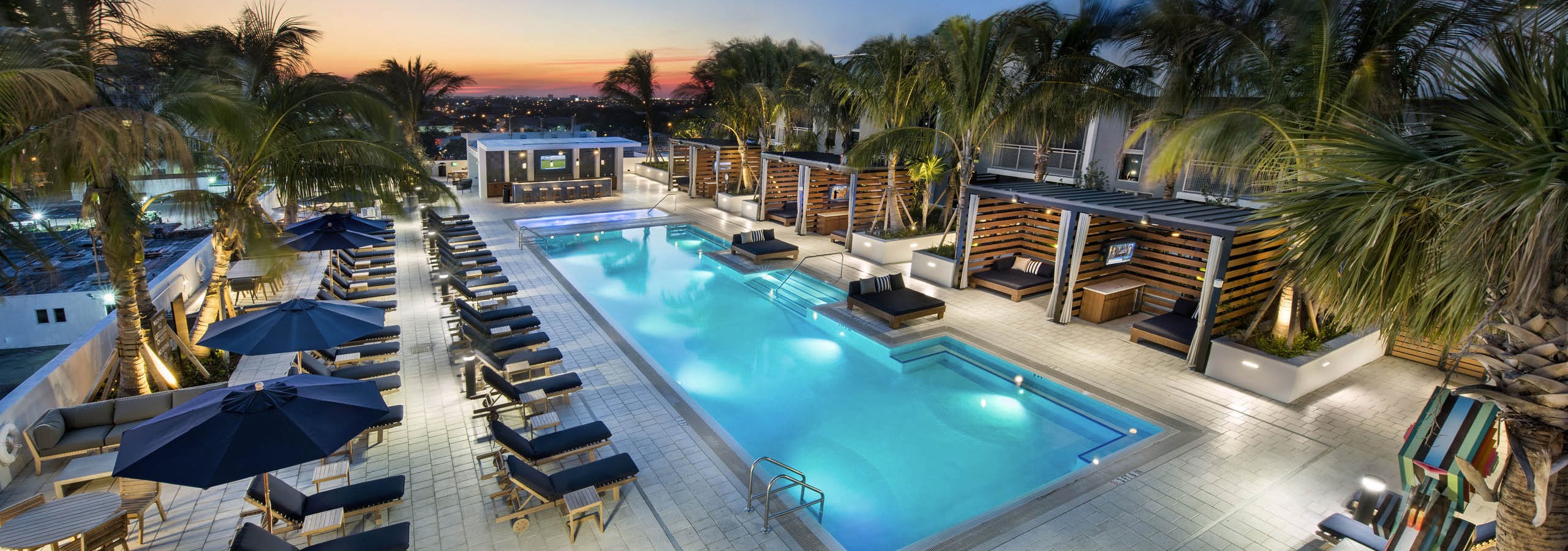 Evening view of AMLI Midtown 29 pool and skyline with individual wood slat cabanas and navy lounge chairs with palm trees