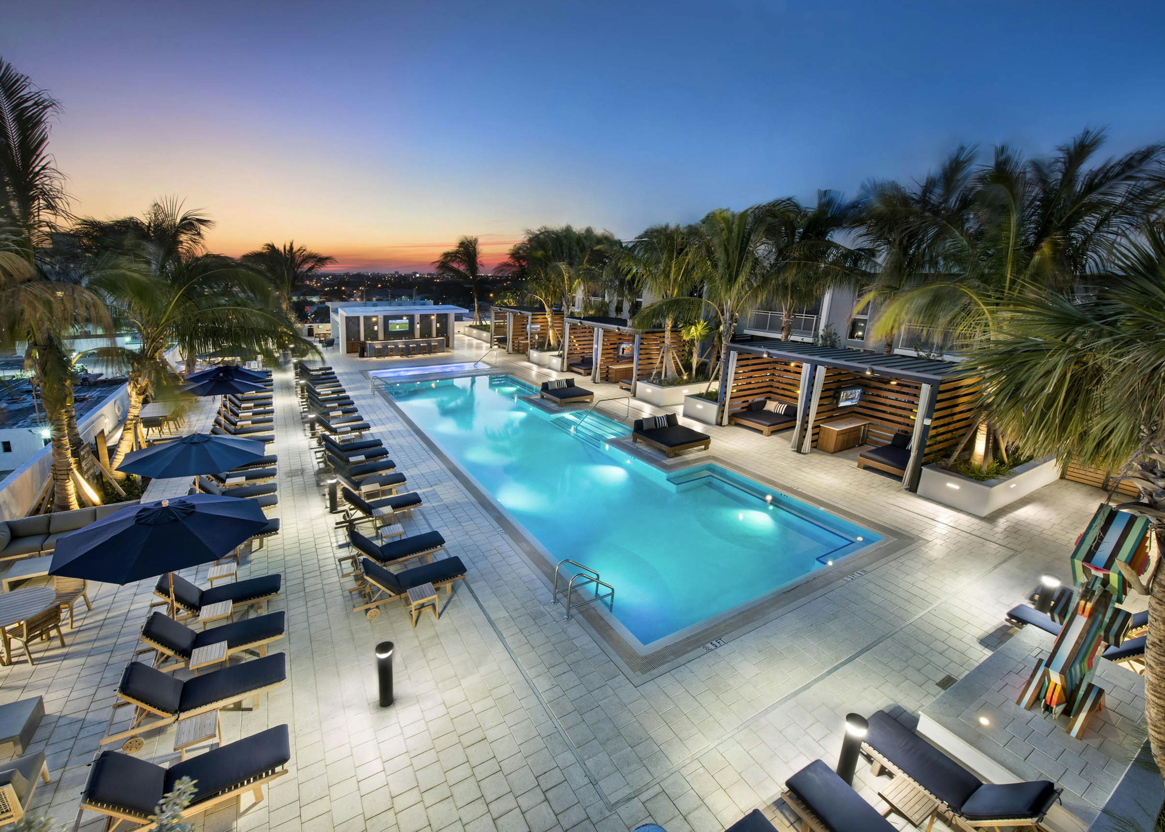 Evening view of AMLI Midtown 29 pool and skyline with individual wood slat cabanas and navy lounge chairs with palm trees