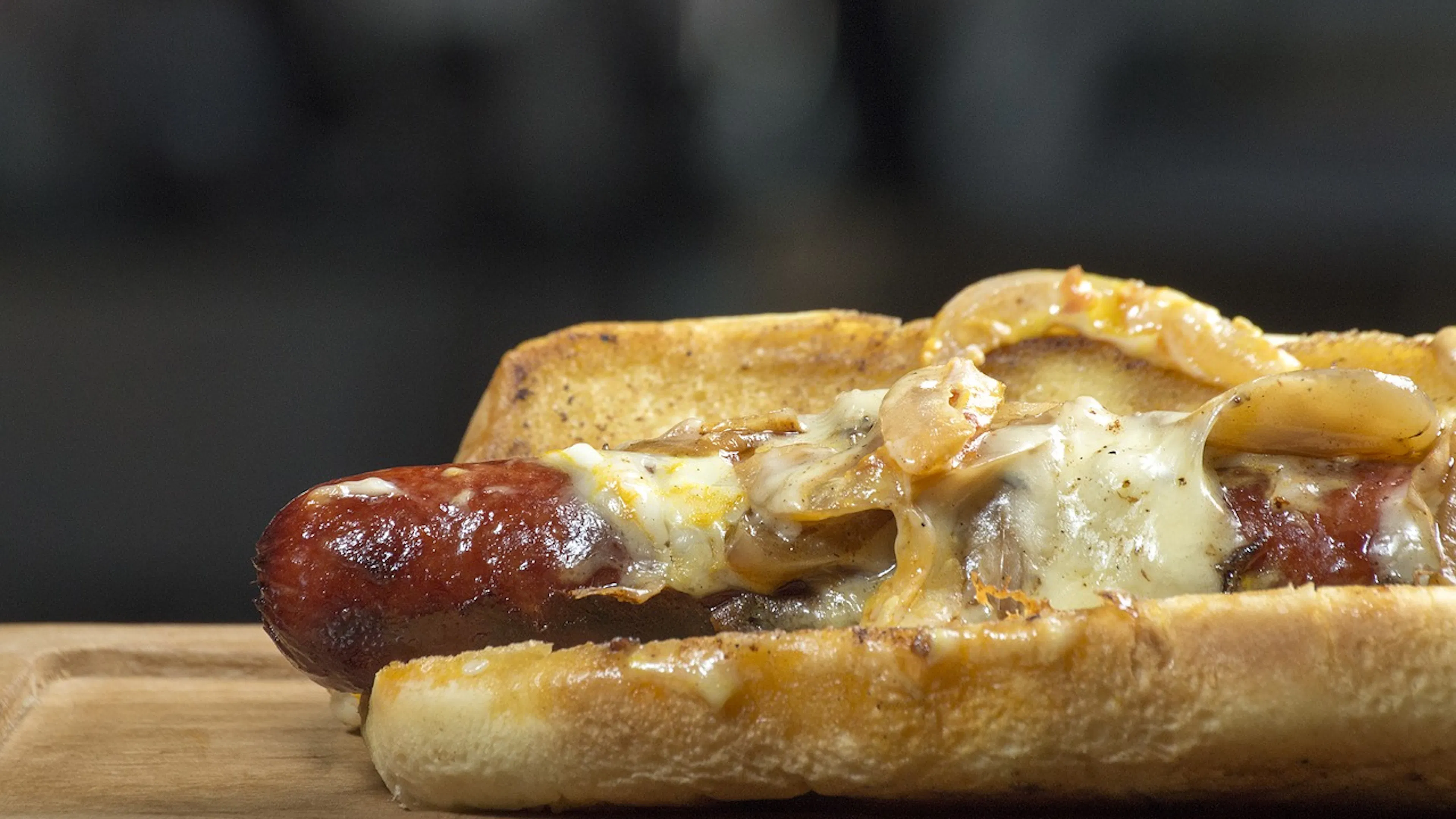 
Close up of a long grilled hot dog on a toasted bun with melted cheddar cheese and sprinkled with sauteed onions