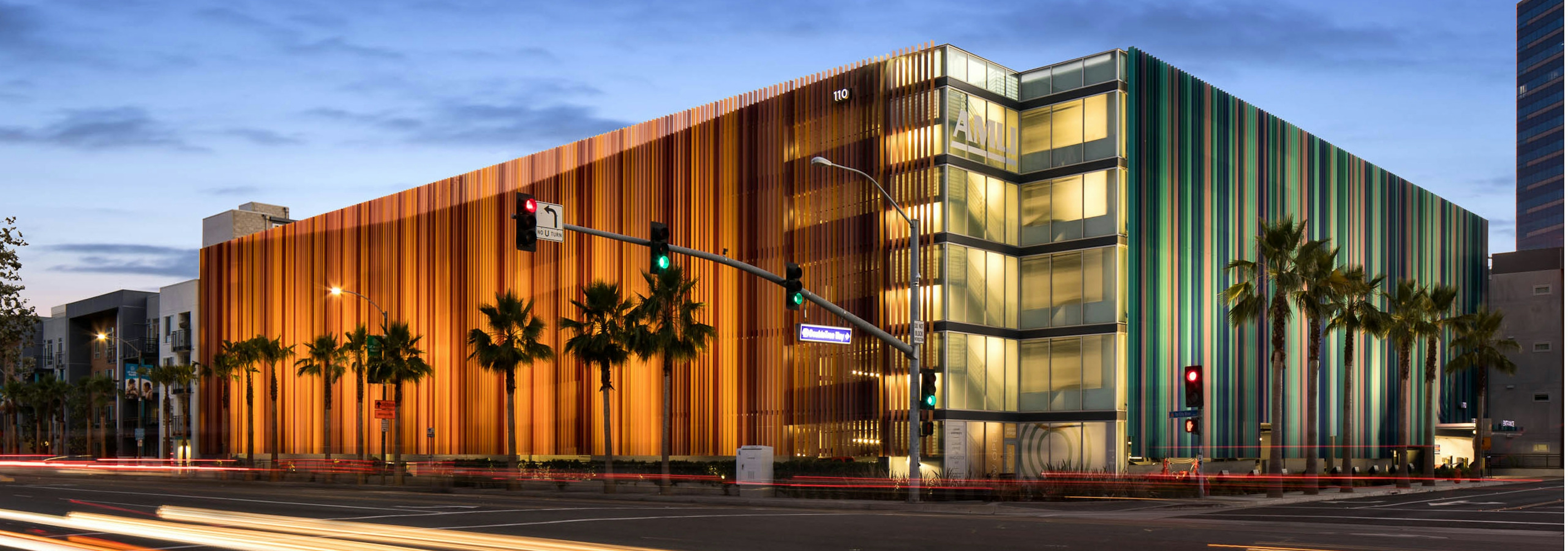 Exterior daytime rendering of AMLI Uptown Orange apartment building rear entrance with lit windows, palm trees and blue sky