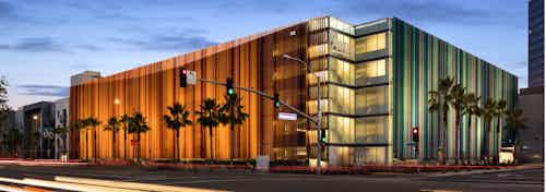 Exterior daytime rendering of AMLI Uptown Orange apartment building rear entrance with lit windows, palm trees and blue sky