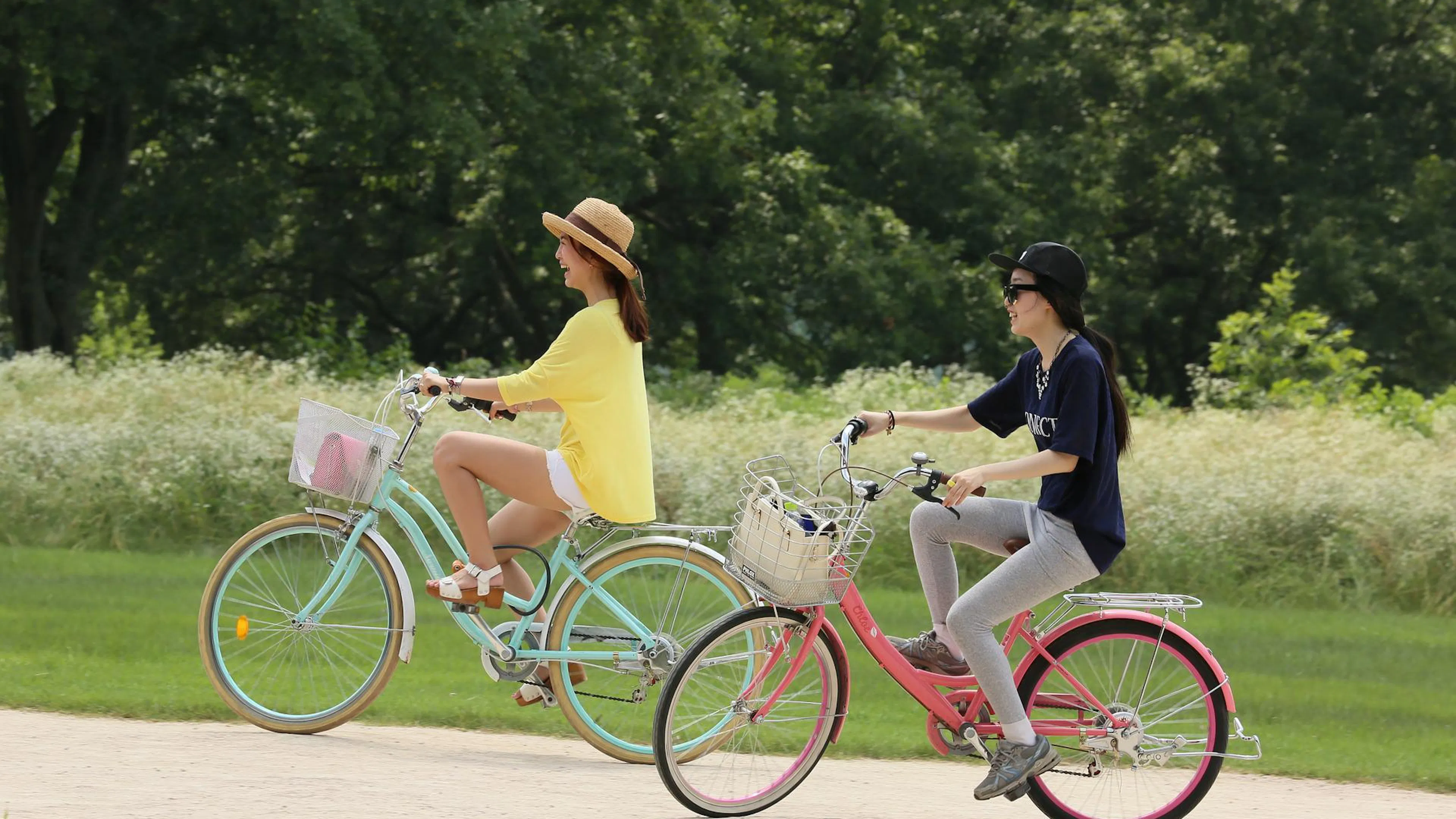 Woman in yellow on blue bike and woman in black on pink bike riding past green trees