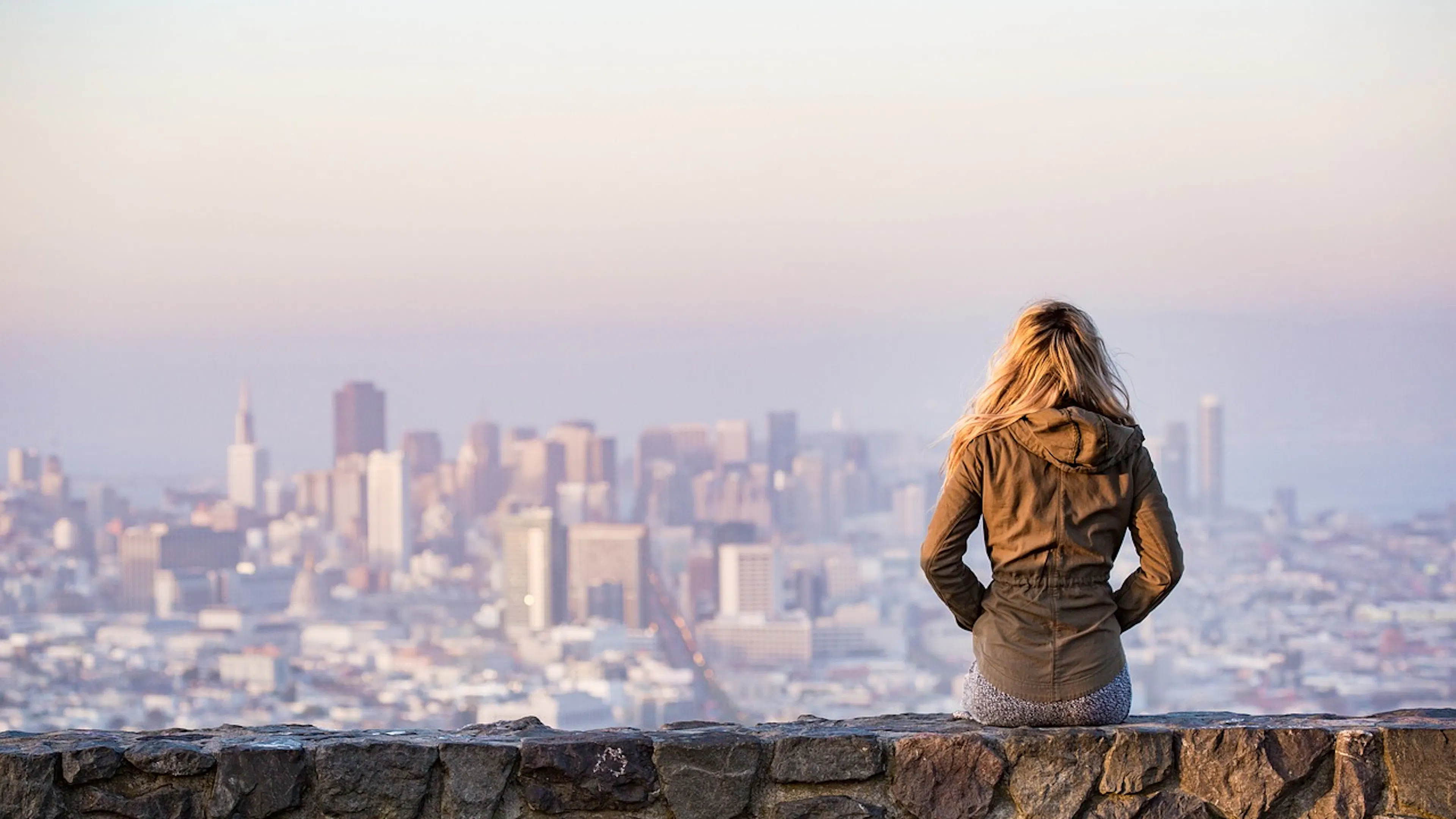 Blonde lady sitting on wall facing away to hazy city skyline