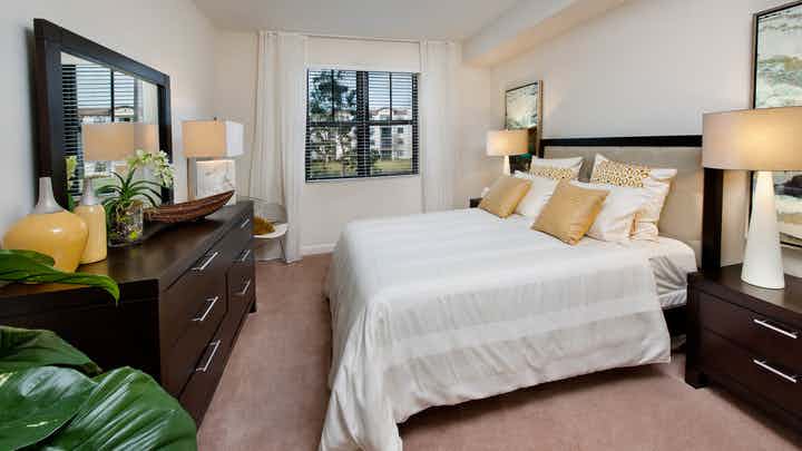 Interior view of an AMLI Doral apartment bedroom with a bed centered on wall, lamps, dresser with mirror and a window view
