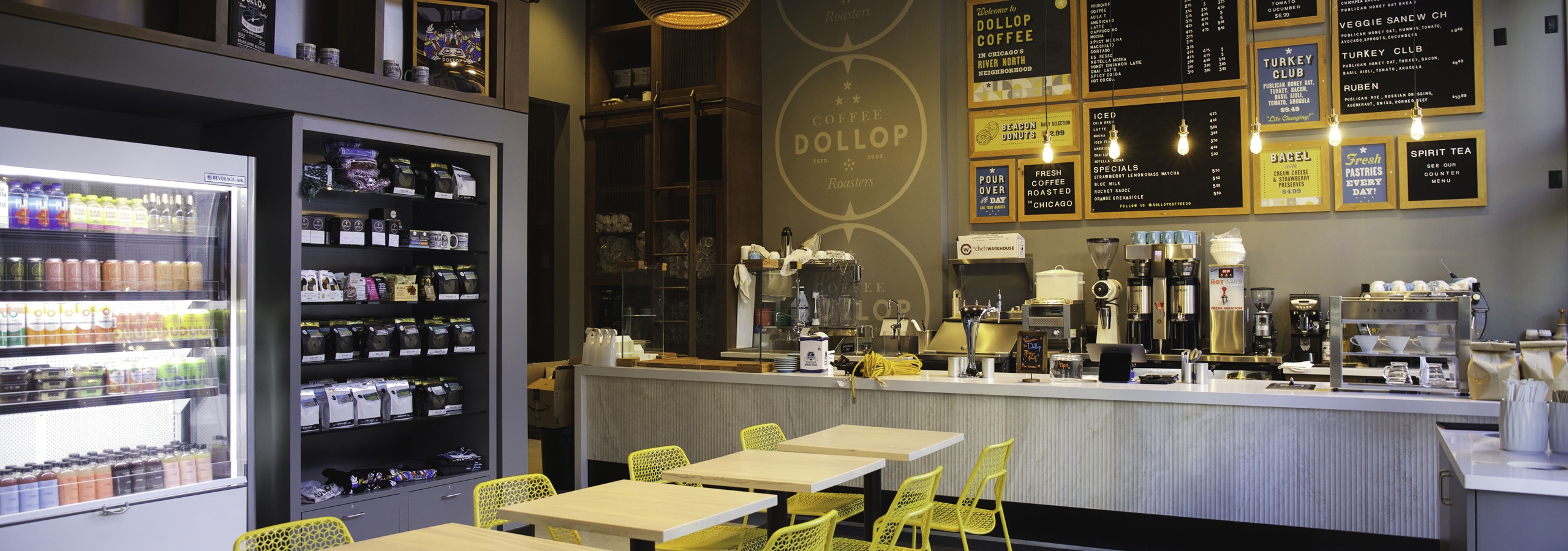 Interior view of Dollop coffee store at AMLI 808 apartment building with 4 tables and 8 yellow chairs and menu boards on wall