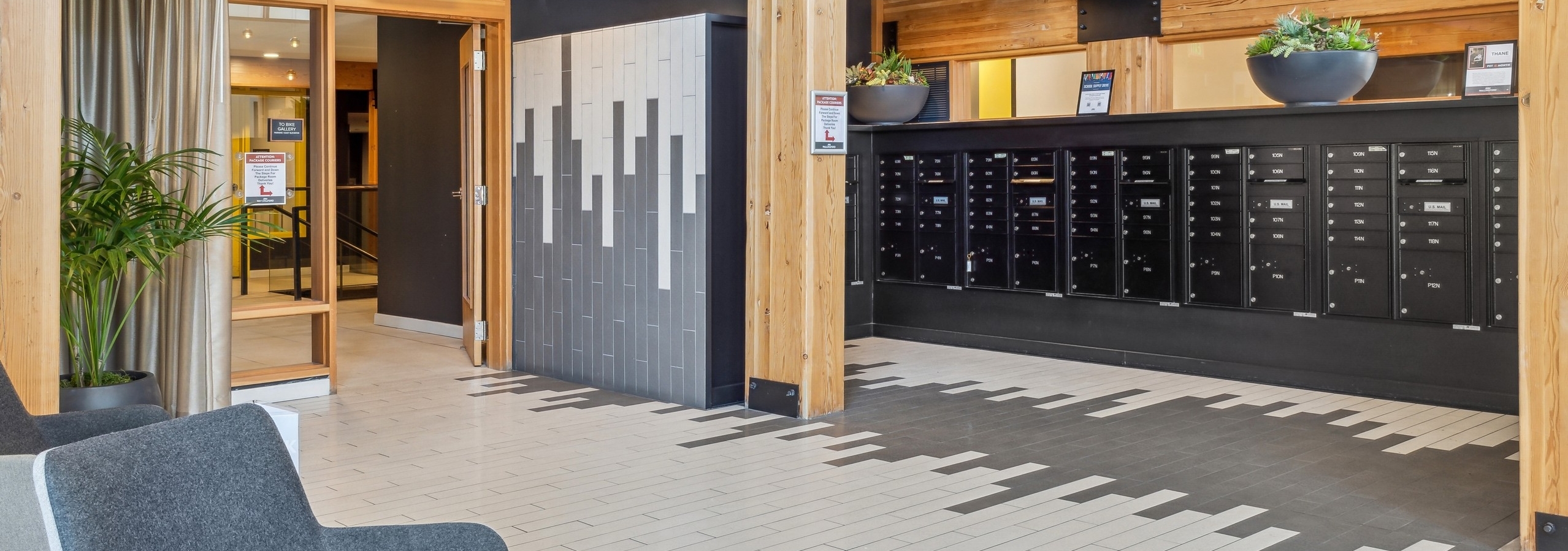 Interior view of a mailroom at AMLI Wallingford with black mailboxes below wood wall and white and grey tile flooring