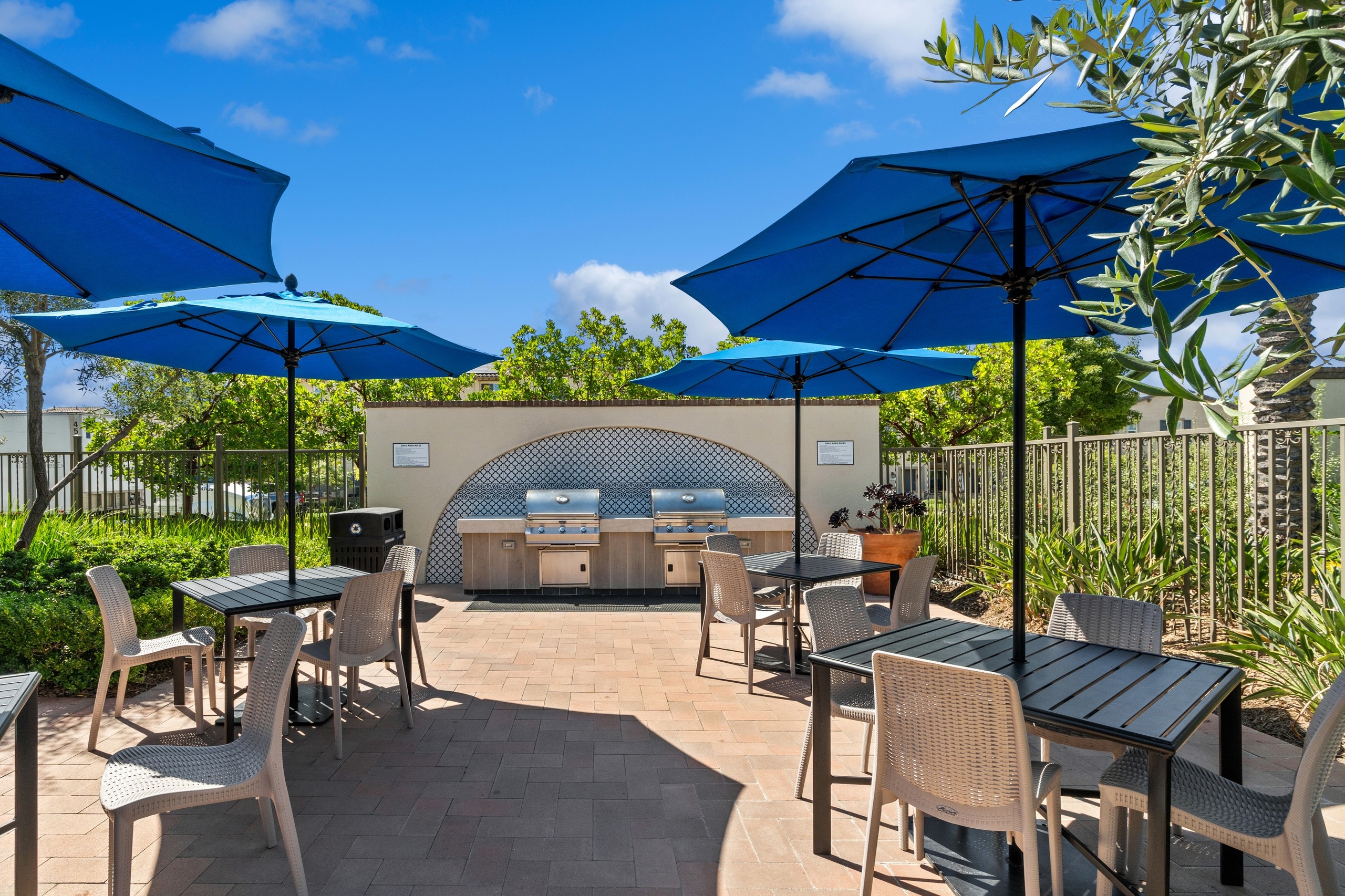 Double grill station in front of tiled wall with 3 square brown tables with chairs and blue umbrellas at AMLI Spanish Hills 