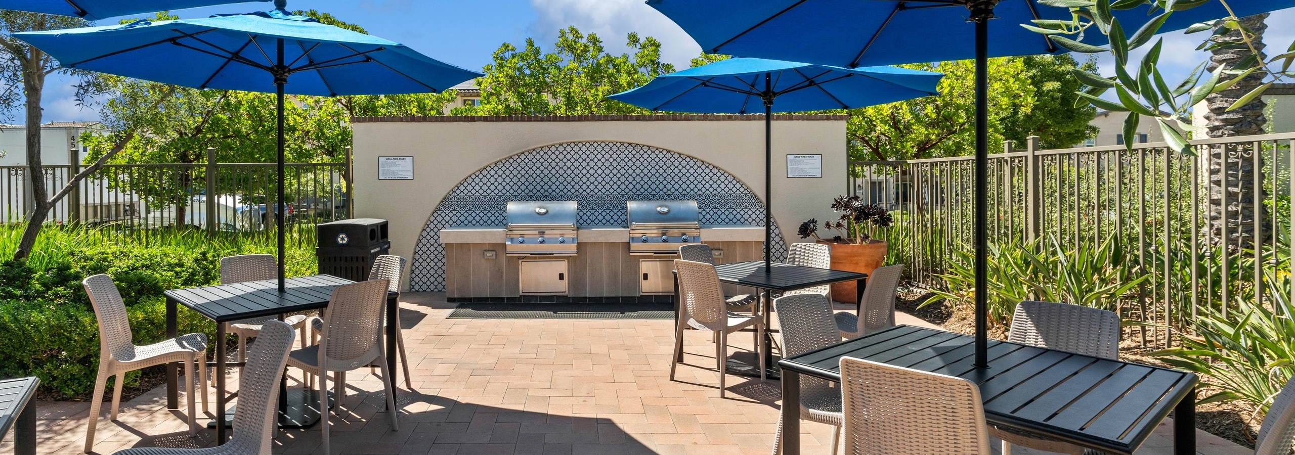 Double grill station in front of tiled wall with 3 square brown tables with chairs and blue umbrellas at AMLI Spanish Hills