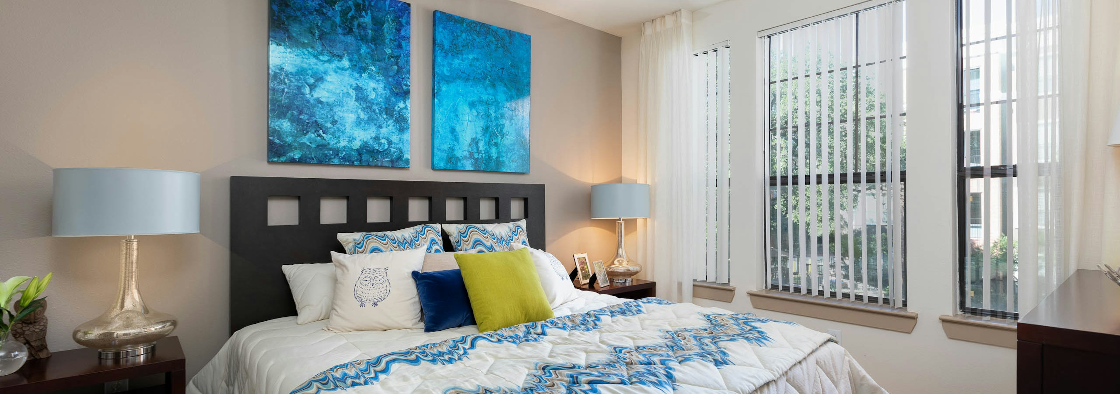 AMLI Eastside bedroom with bright windows and vibrant blue wall art hanging on a tan accent wall above a white bed