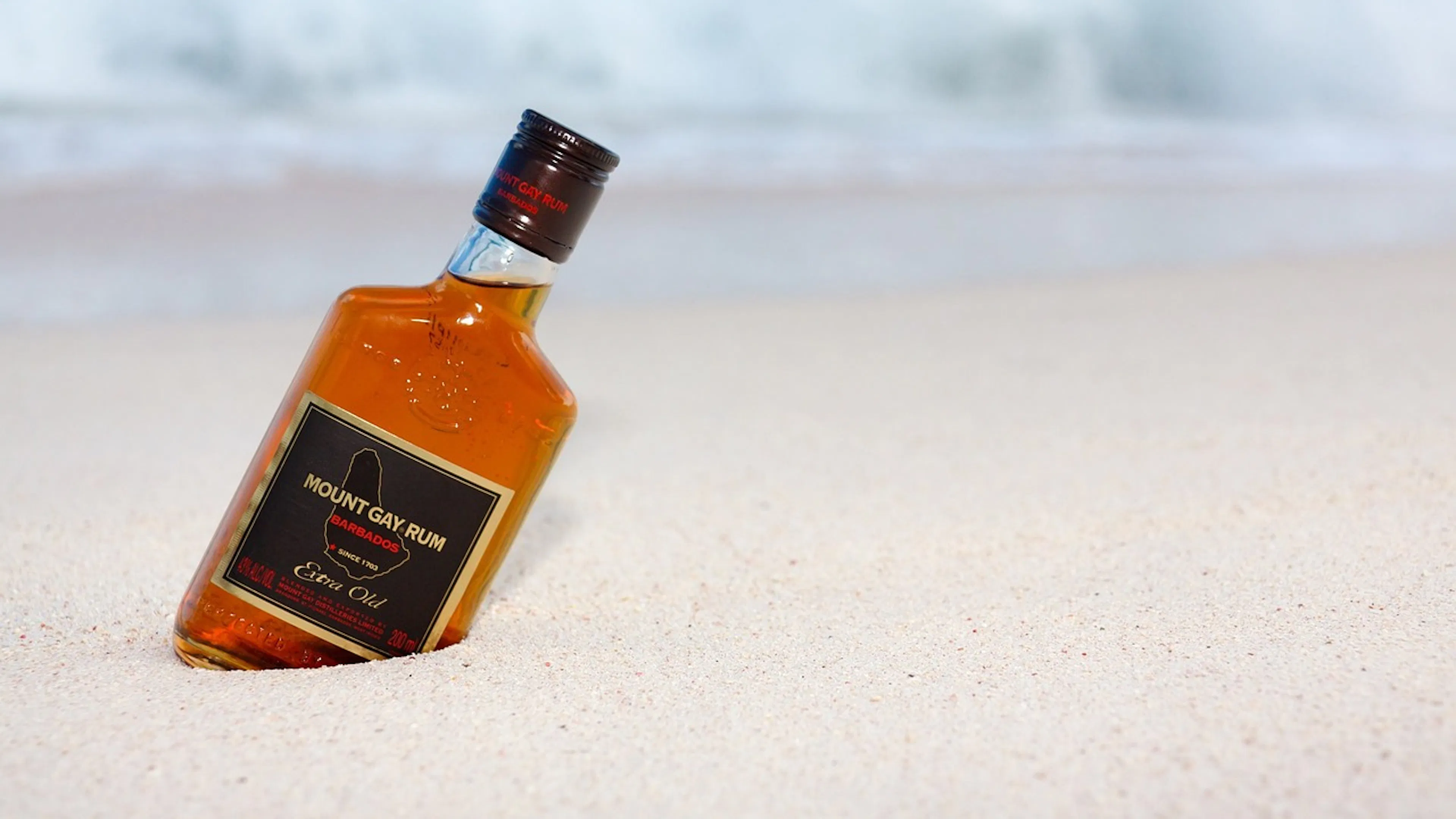 A bottle of gold rum lying crooked in white sand with waves crashing in the background