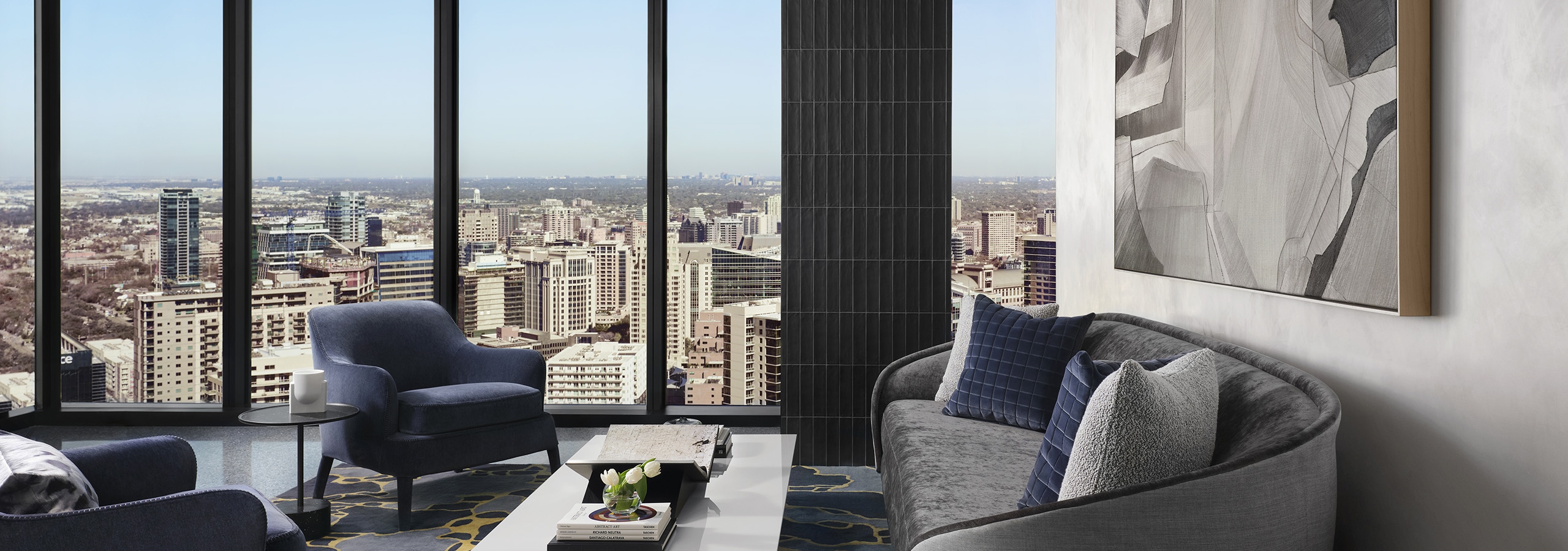 Resident lounge with a plush gray couch and two blue chairs around a coffee table facing expansive skyline view