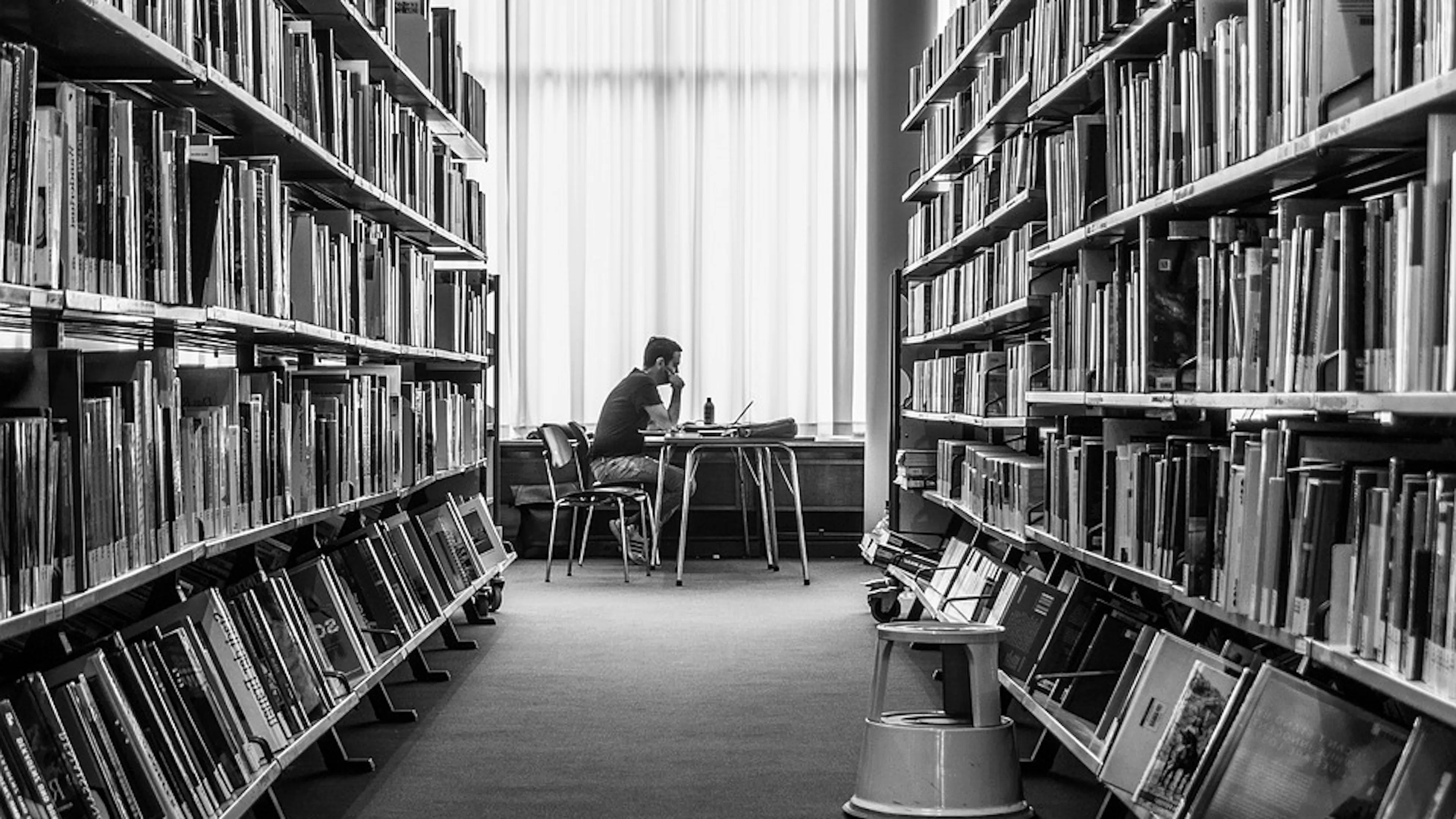 A black and white picture of a man sitting at a table reading a book at the end of an aisle of books