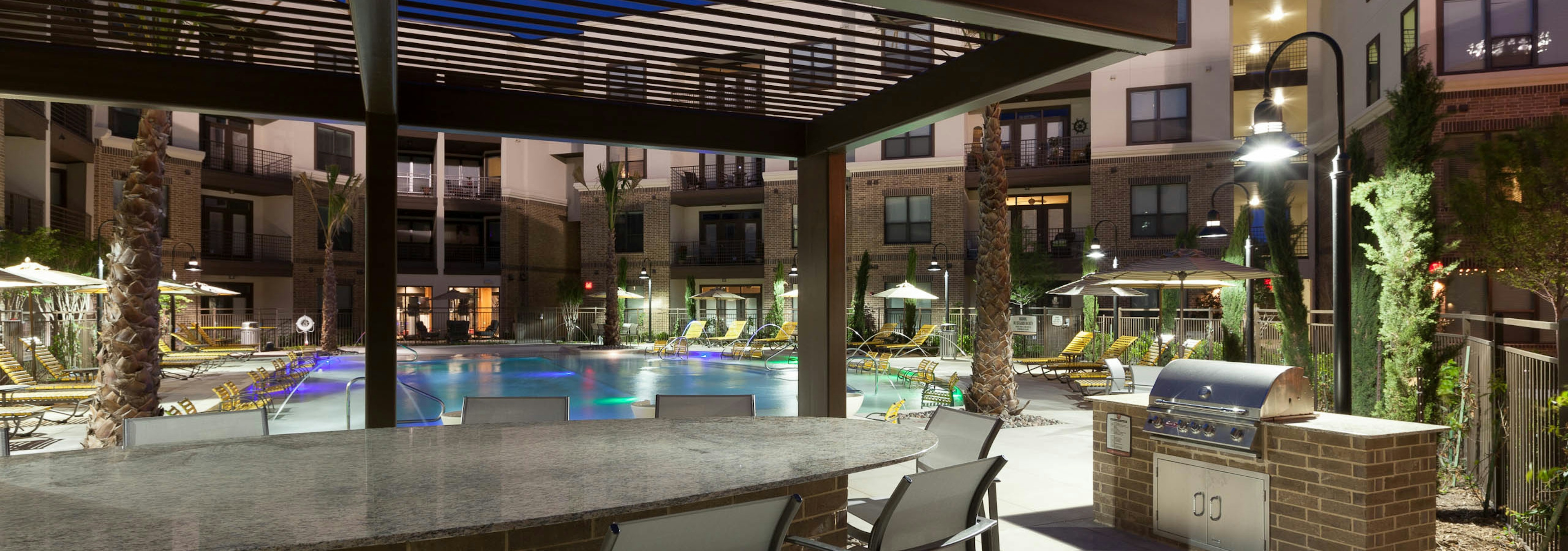 Nighttime outdoor view of grill station, seating and pool area at AMLI at the Ballpark apartments with buildings in background