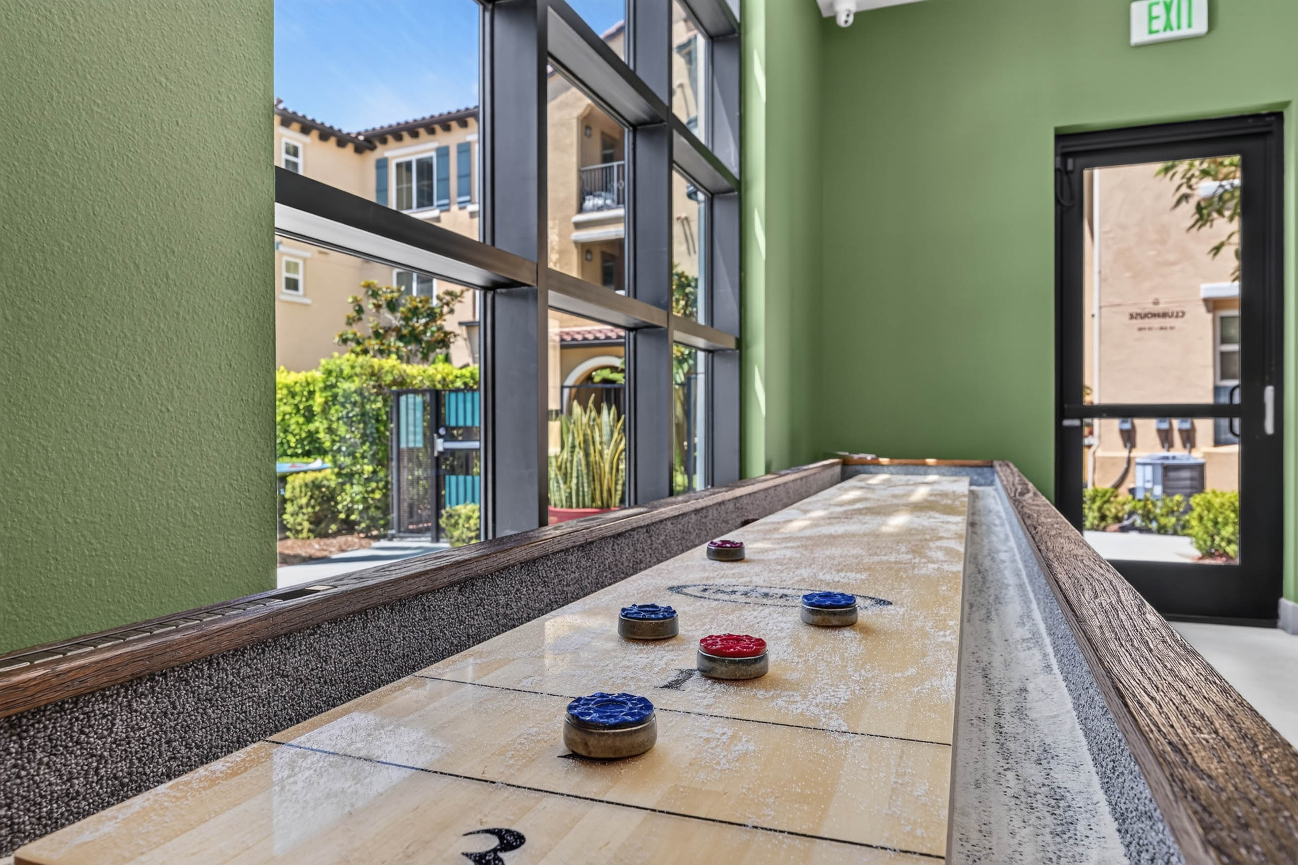 Shuffleboard table at Game Terrace with green walls and large windows overlooking landscaped grounds at AMLI Camarillo apartments.